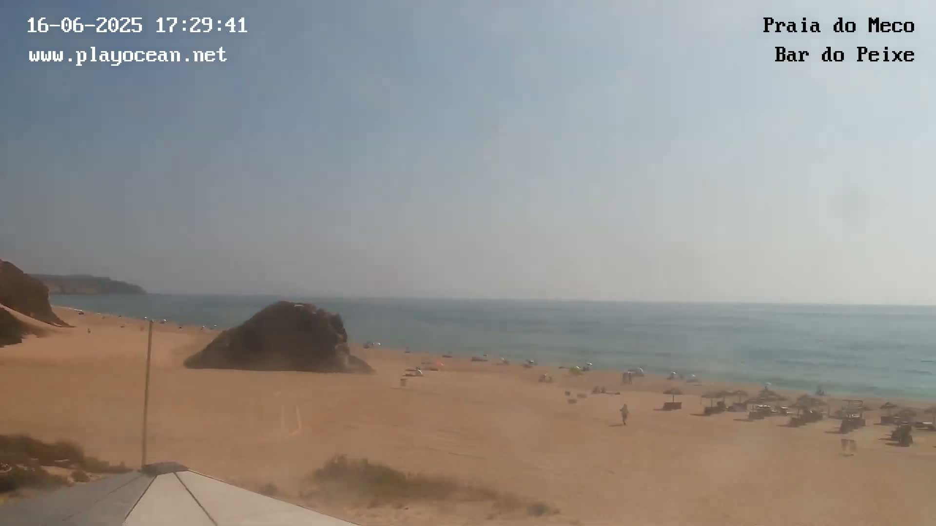 A sandy beach with scattered people and beach umbrellas stretches to a calm ocean under a clear, bright sky.