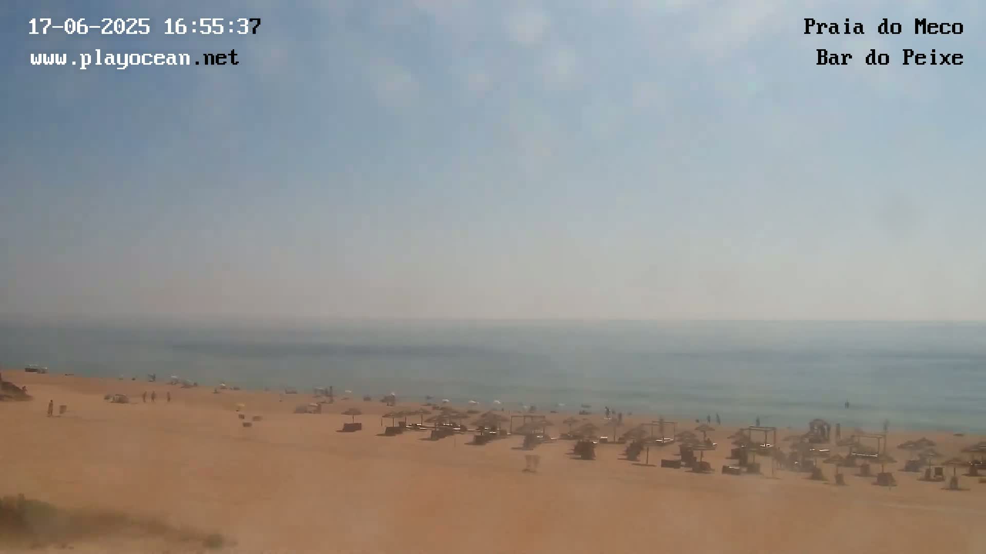 A hazy, sunny day at a beach shows a mostly empty sandy shore with a few people and beach umbrellas, and a calm ocean in the background.