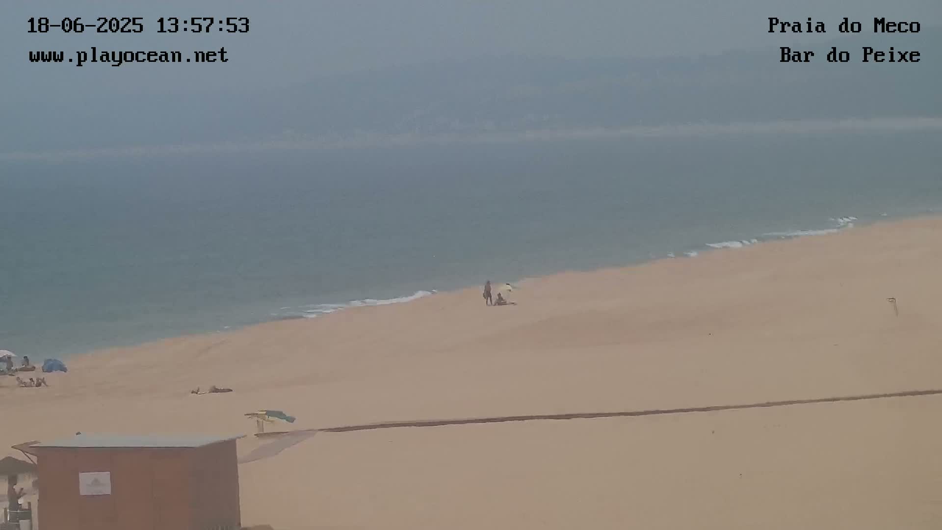 A hazy view shows a wide sandy beach with a few people and a small structure, meeting a calm, light-blue ocean.