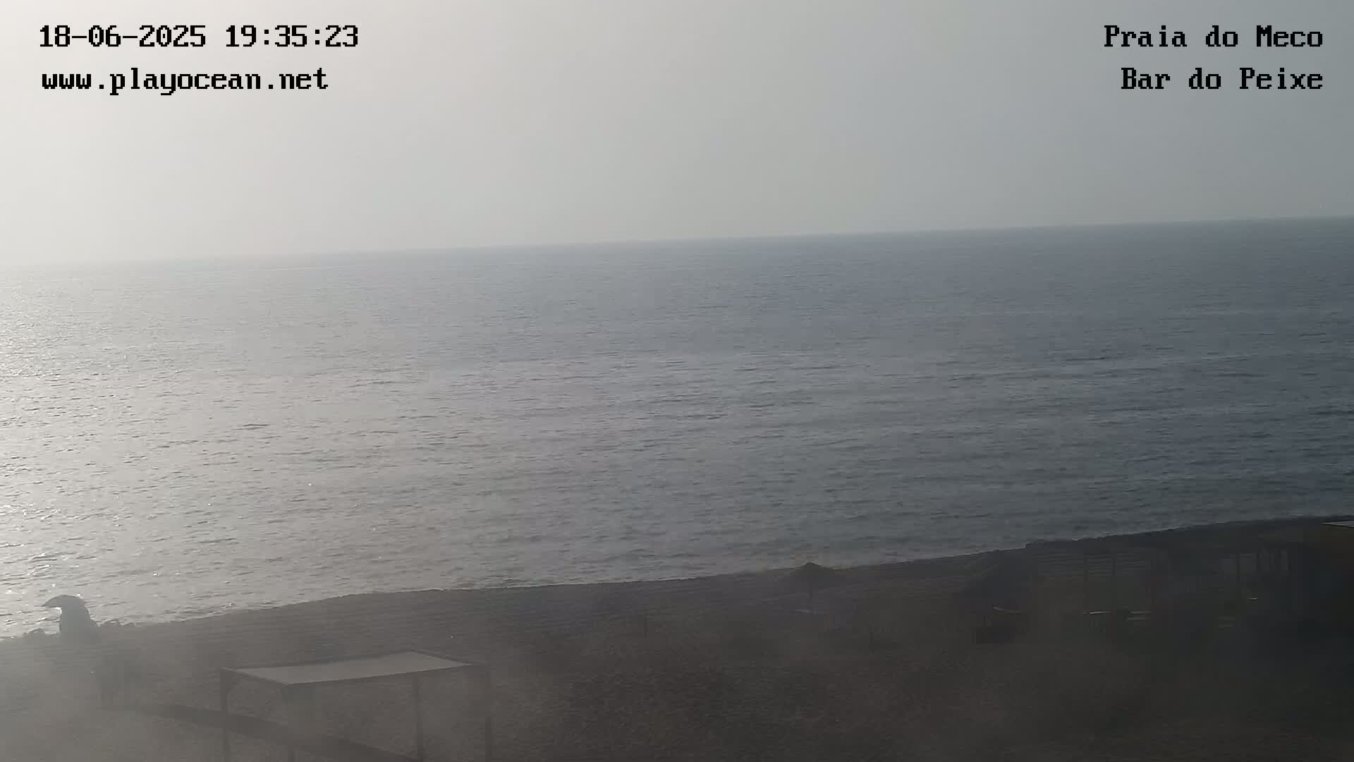 A hazy view shows a calm ocean meeting a dark sandy beach with some indistinct structures.
