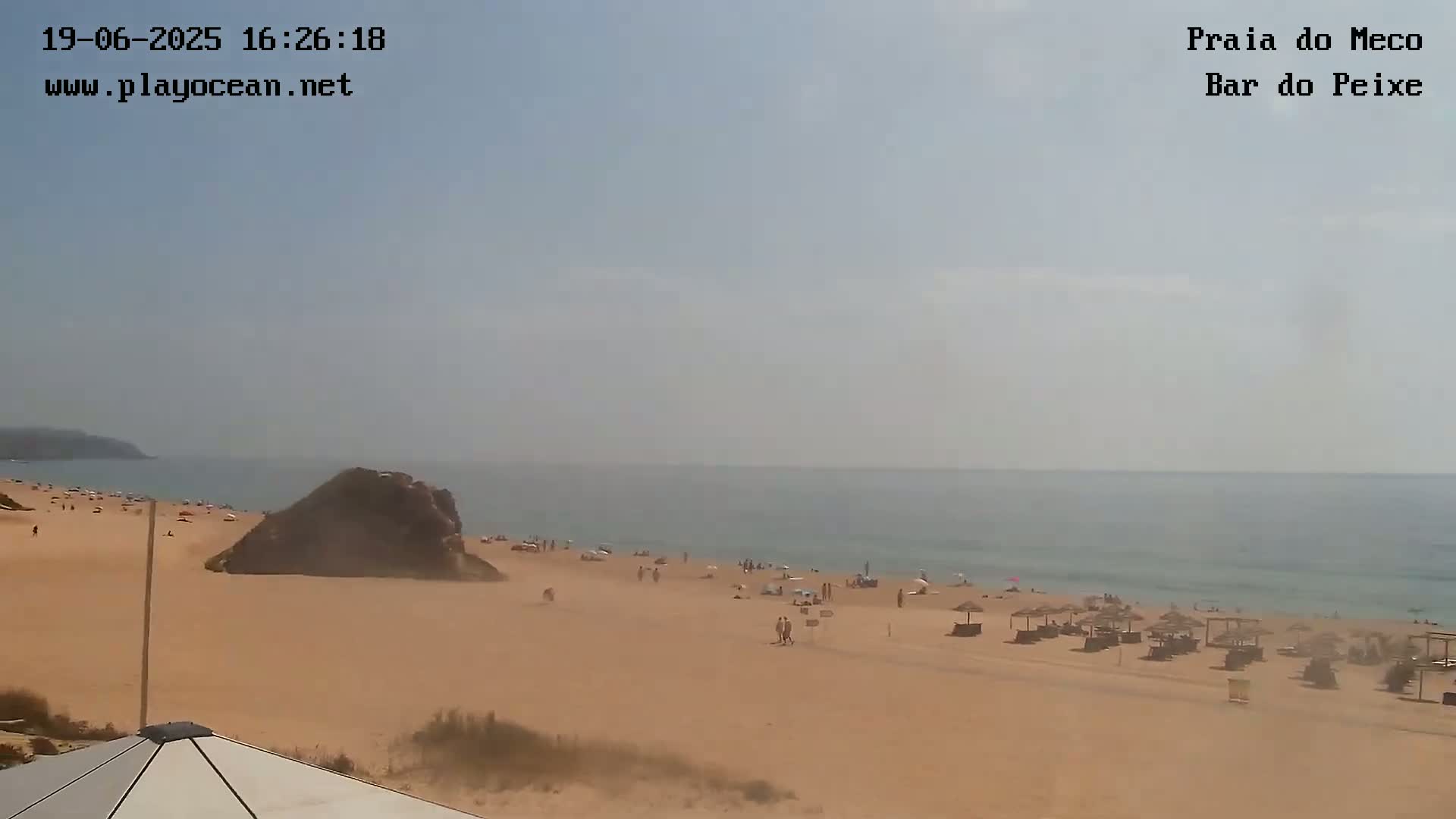 A sandy beach with scattered people and beach umbrellas stretches along a calm, hazy ocean under a mostly clear sky.