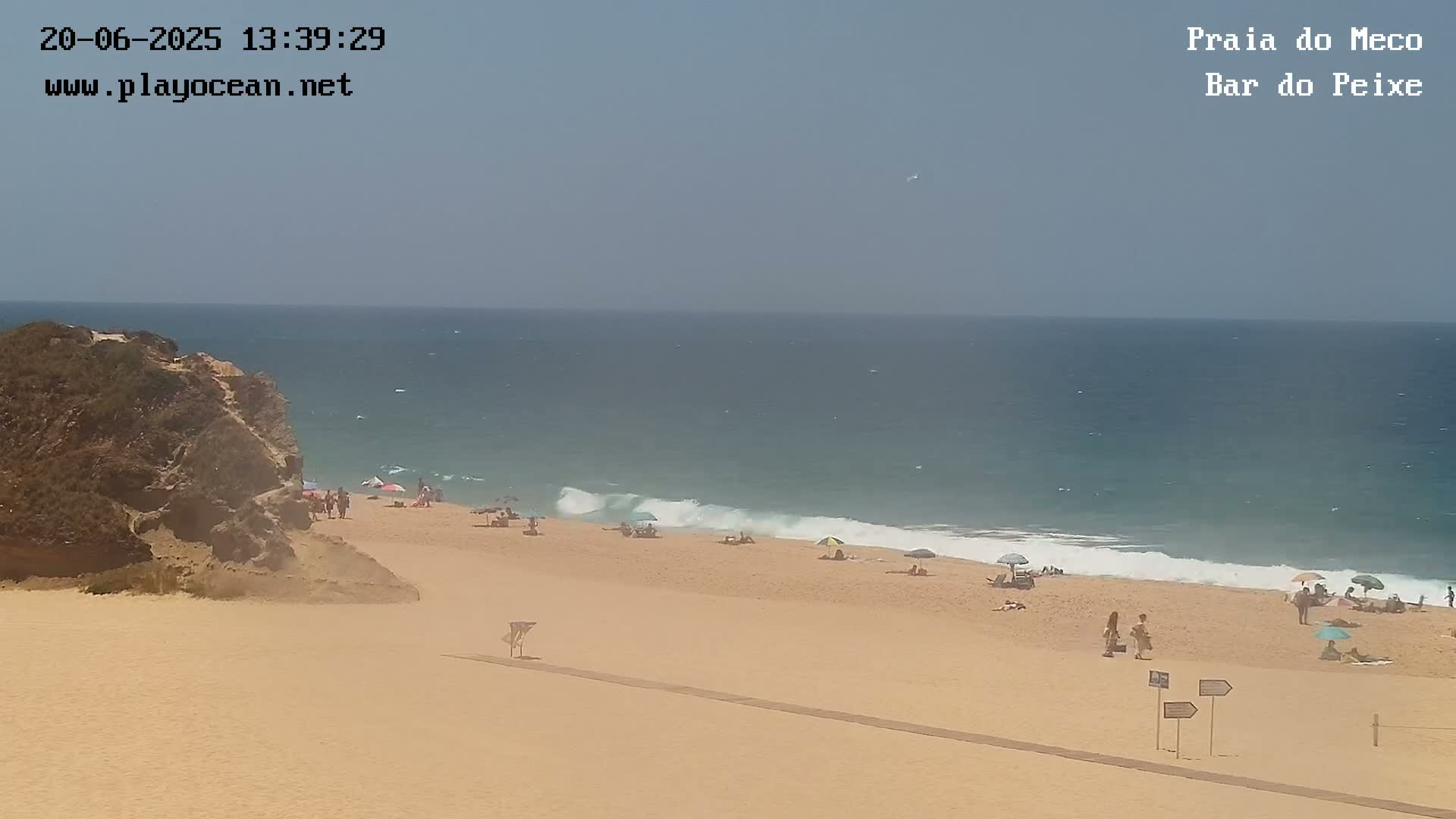 A sandy beach with a few people and umbrellas under a clear, sunny sky, next to a calm ocean with small waves breaking on the shore and a rocky outcrop to the left.