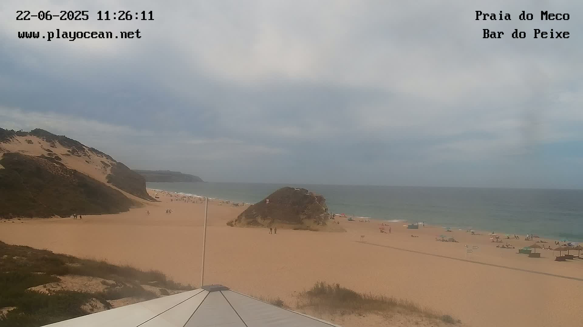 A sandy beach with a few people on it, backed by sand dunes and a calm ocean under a mostly cloudy sky.