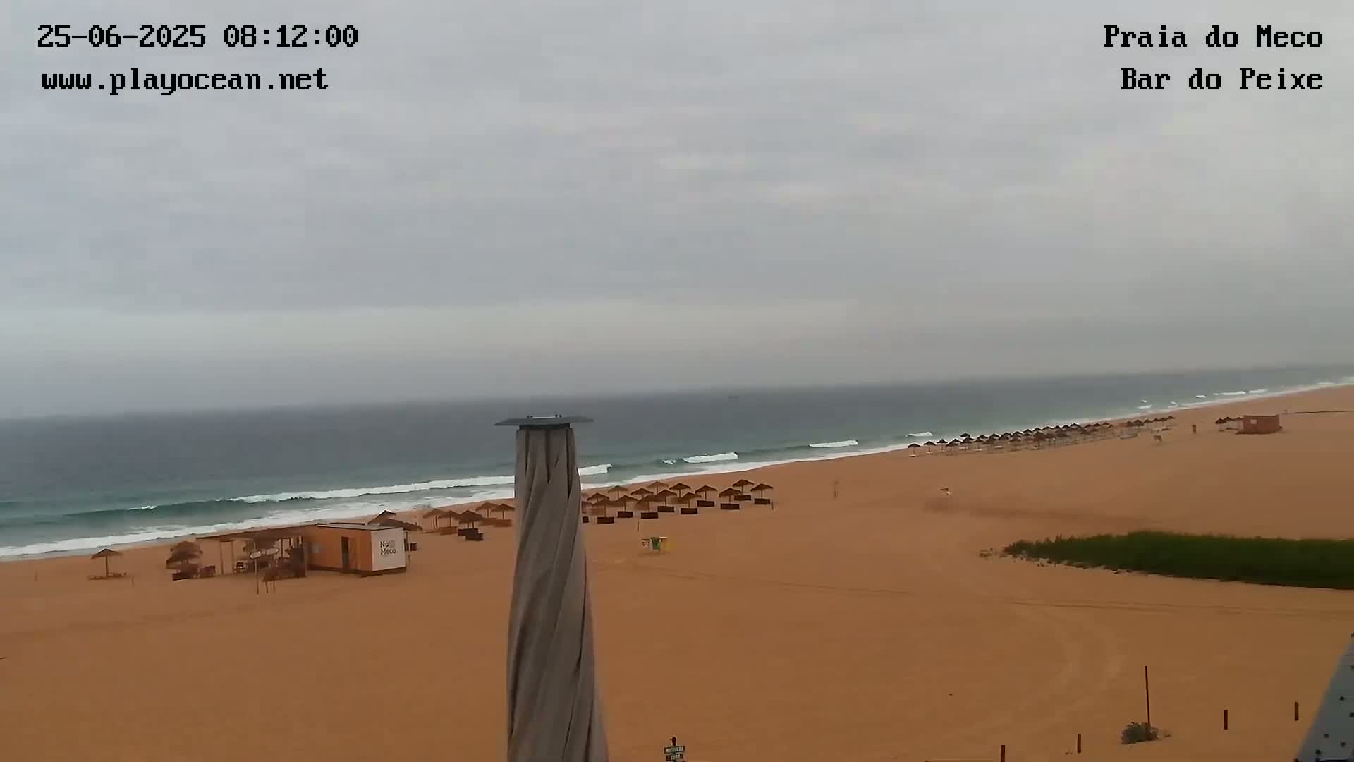 A mostly empty sandy beach with a few umbrellas and a small building is seen under an overcast sky, with gentle waves rolling in on a calm ocean.
