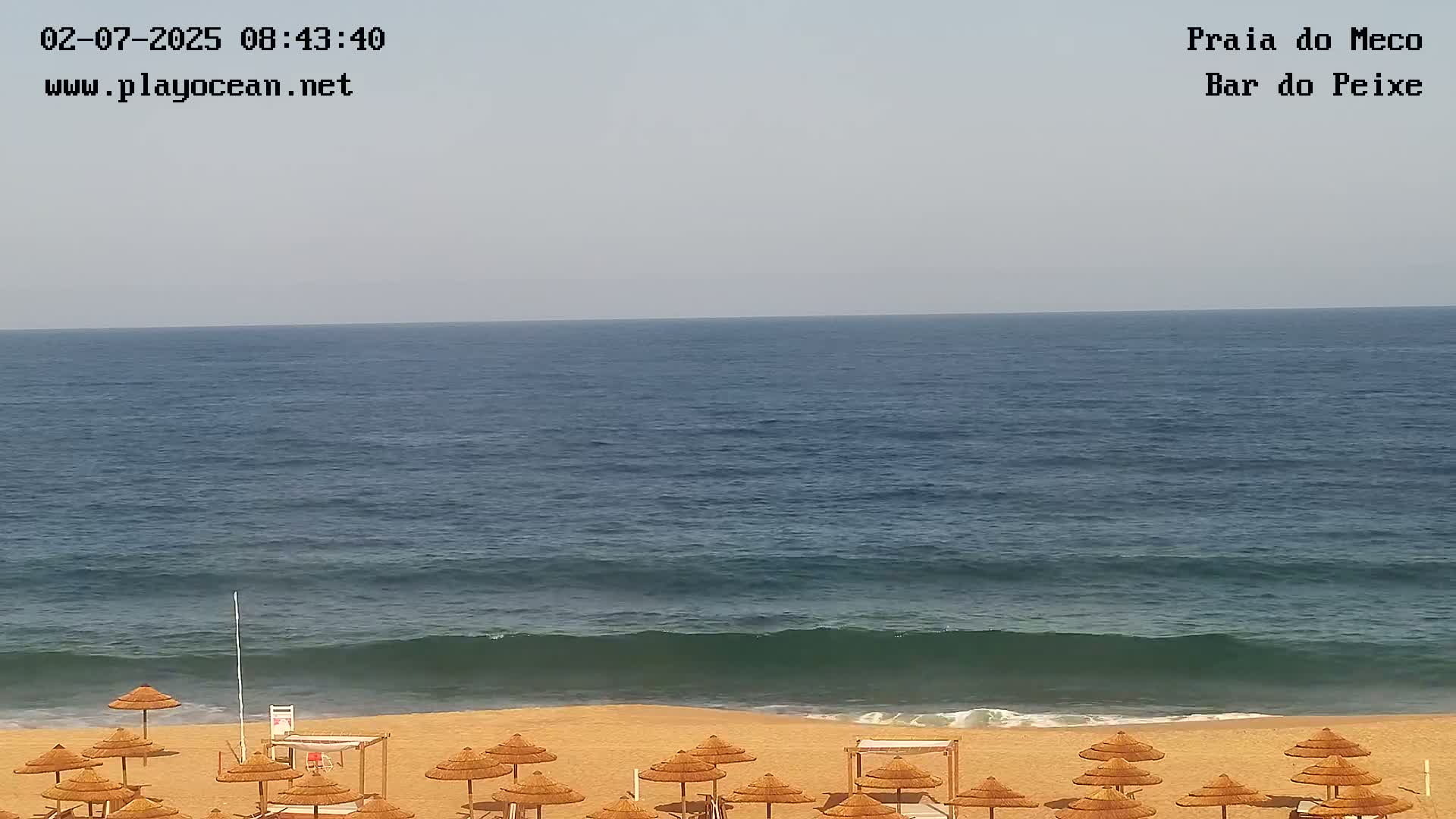 A sandy beach with straw umbrellas is shown, overlooking a calm ocean under a clear sky.