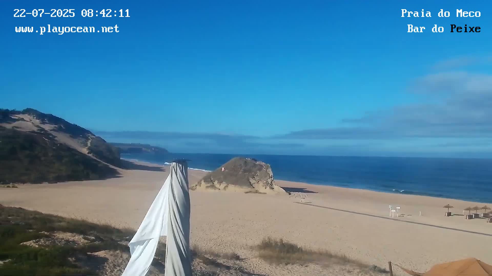 A sandy beach with calm ocean waves stretches beneath a clear blue sky with a few scattered clouds, and a large sand dune is visible to the left.