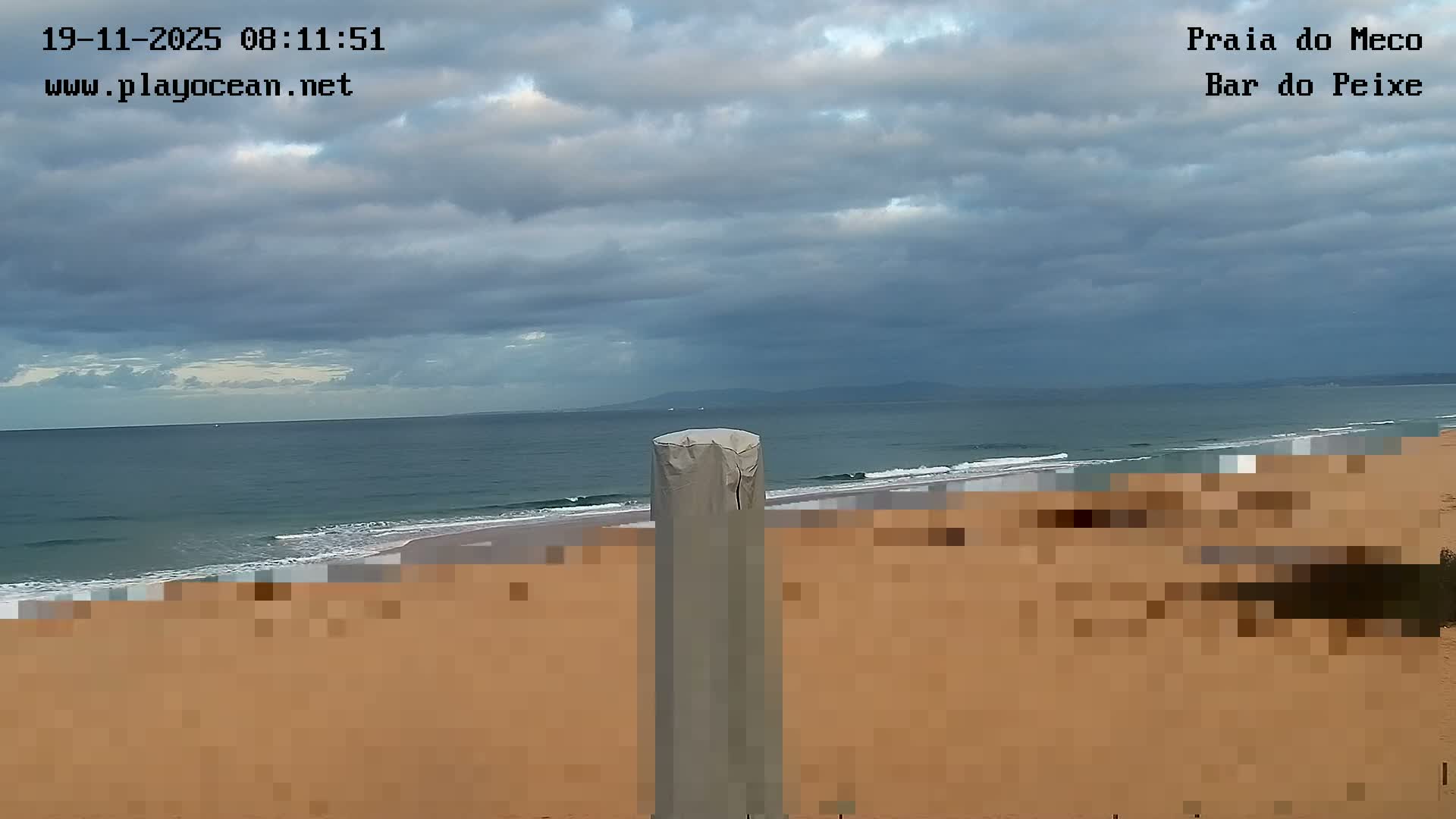 Heavily overcast and cloudy skies hang above a blue-green ocean with breaking waves, meeting a pixelated sandy beach with a gray covered pole in the foreground and distant land on the horizon.
