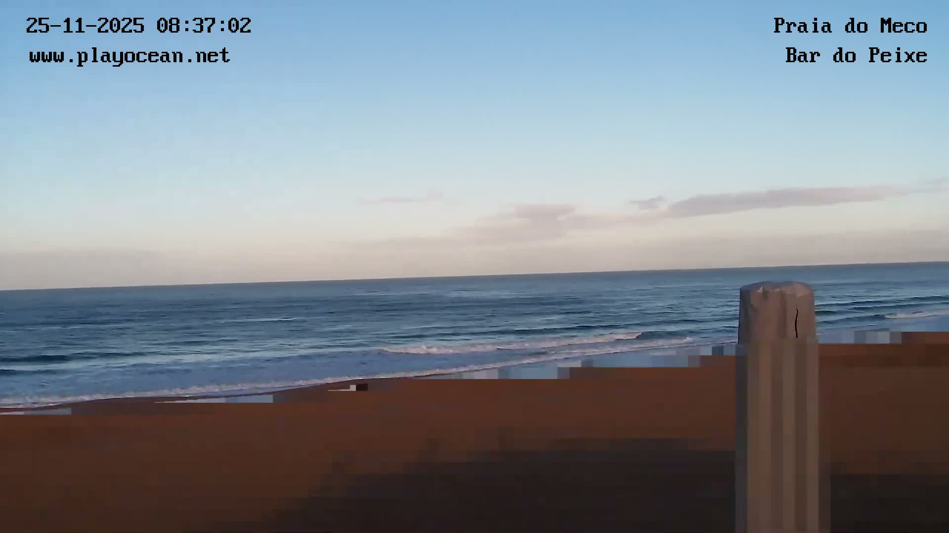 A mostly clear, light blue sky with faint clouds overlooks a blue ocean with breaking waves and a dark sandy foreground, partially obstructed by a grey post on the right.