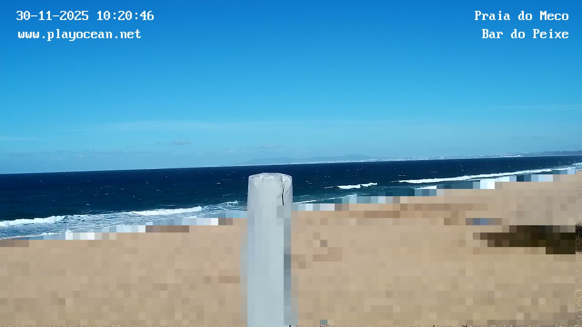 A clear, sunny day overlooks a wide sandy beach with dark blue ocean waves crashing onto the shore, while distant land masses are visible under a bright blue sky, partially obscured by a white pole in the foreground.