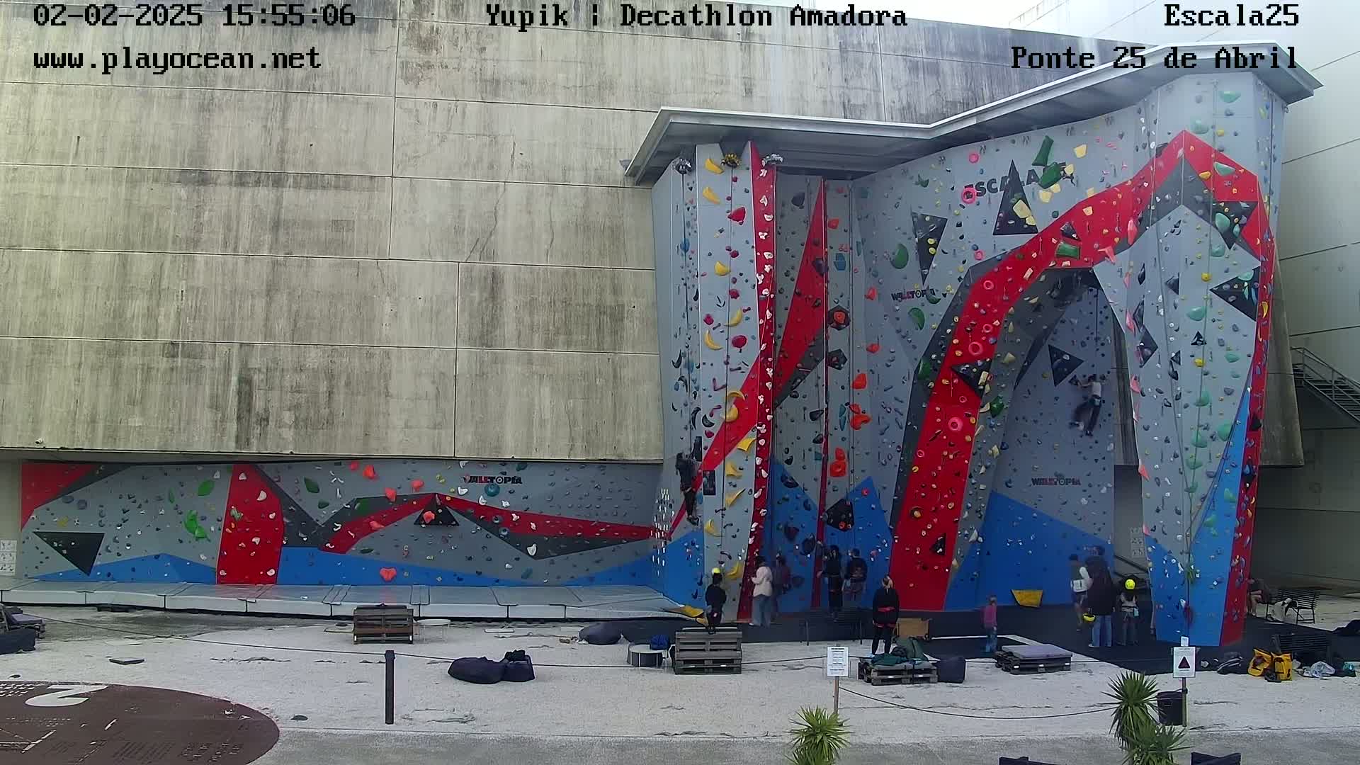 A large outdoor climbing wall with various colored holds, and several people climbing or watching from the ground on a mostly cloudy day.