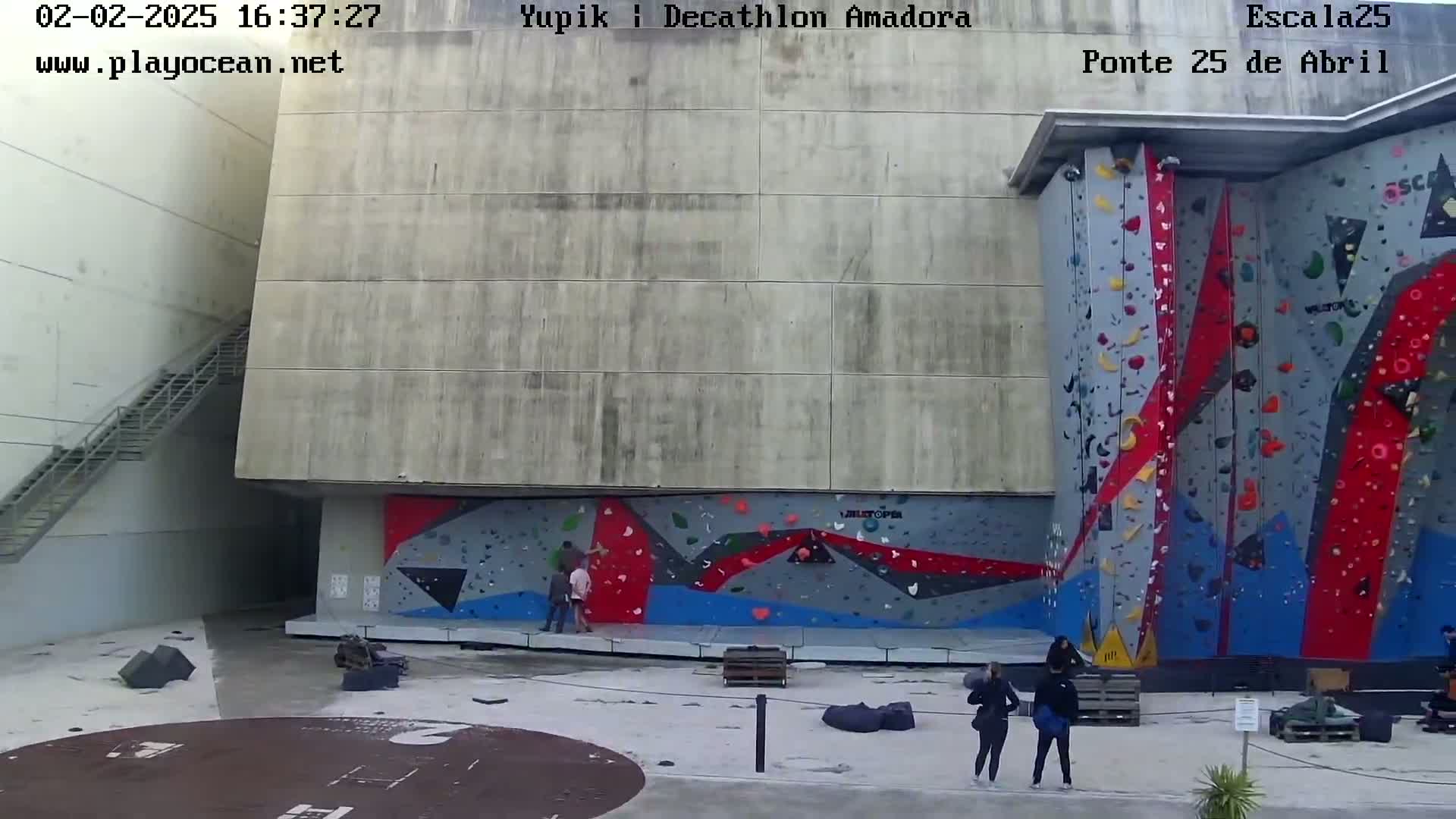 An outdoor scene shows a large, multi-colored climbing wall next to a concrete building, with a few people standing nearby on a mostly clear day.