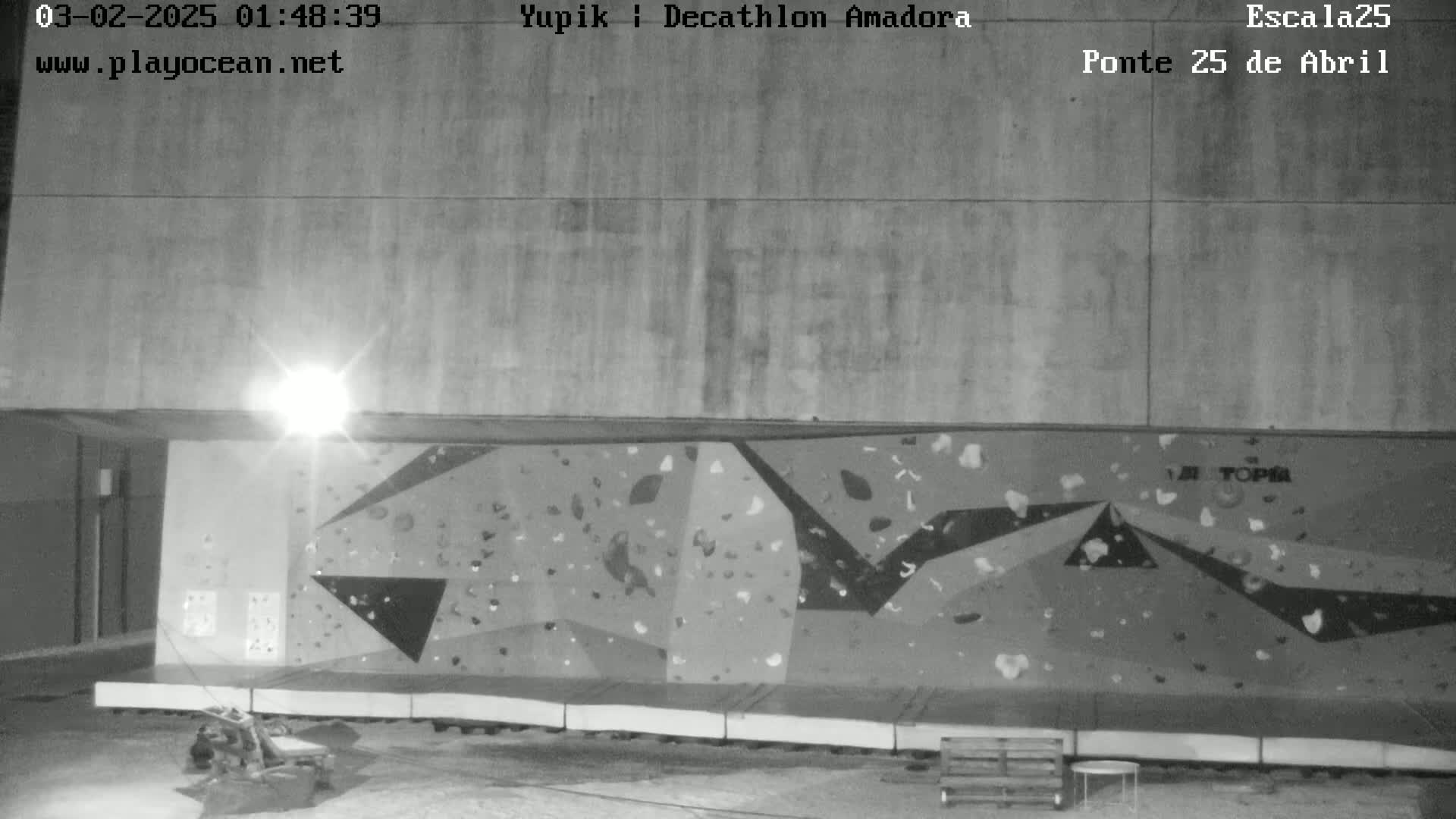 A nighttime grayscale image shows an outdoor climbing wall beneath a concrete overhang, illuminated by a bright light source.