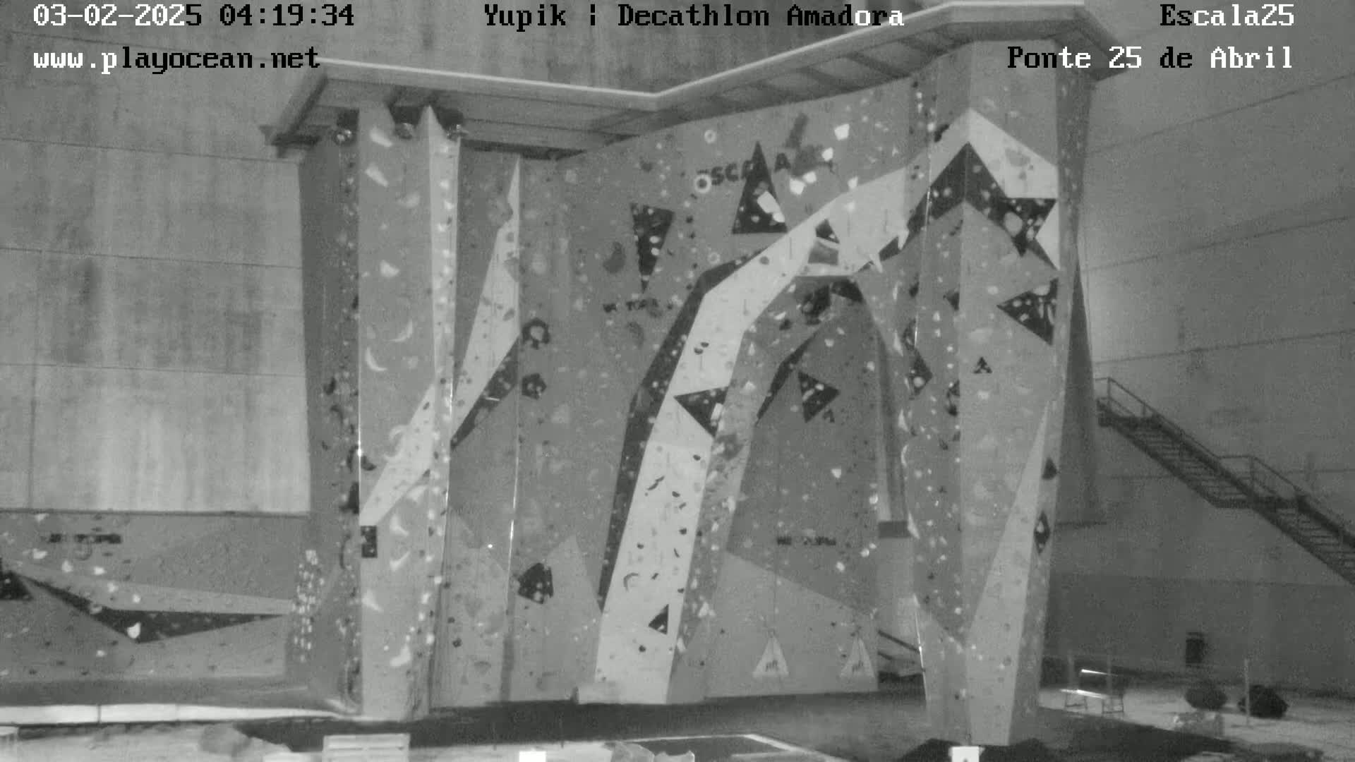 A large indoor climbing wall with various holds and patterns stands in a gray, dimly lit building.