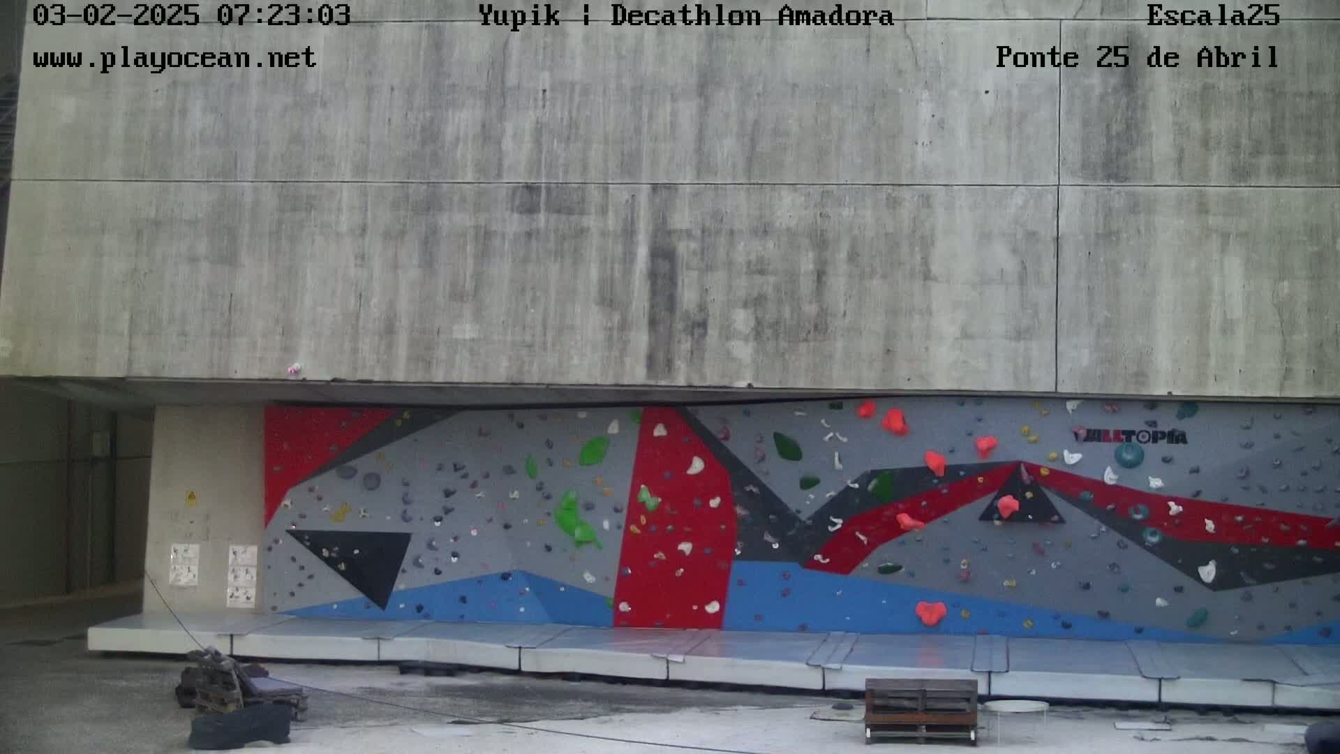 An outdoor climbing wall with various colored holds is situated against a concrete building.