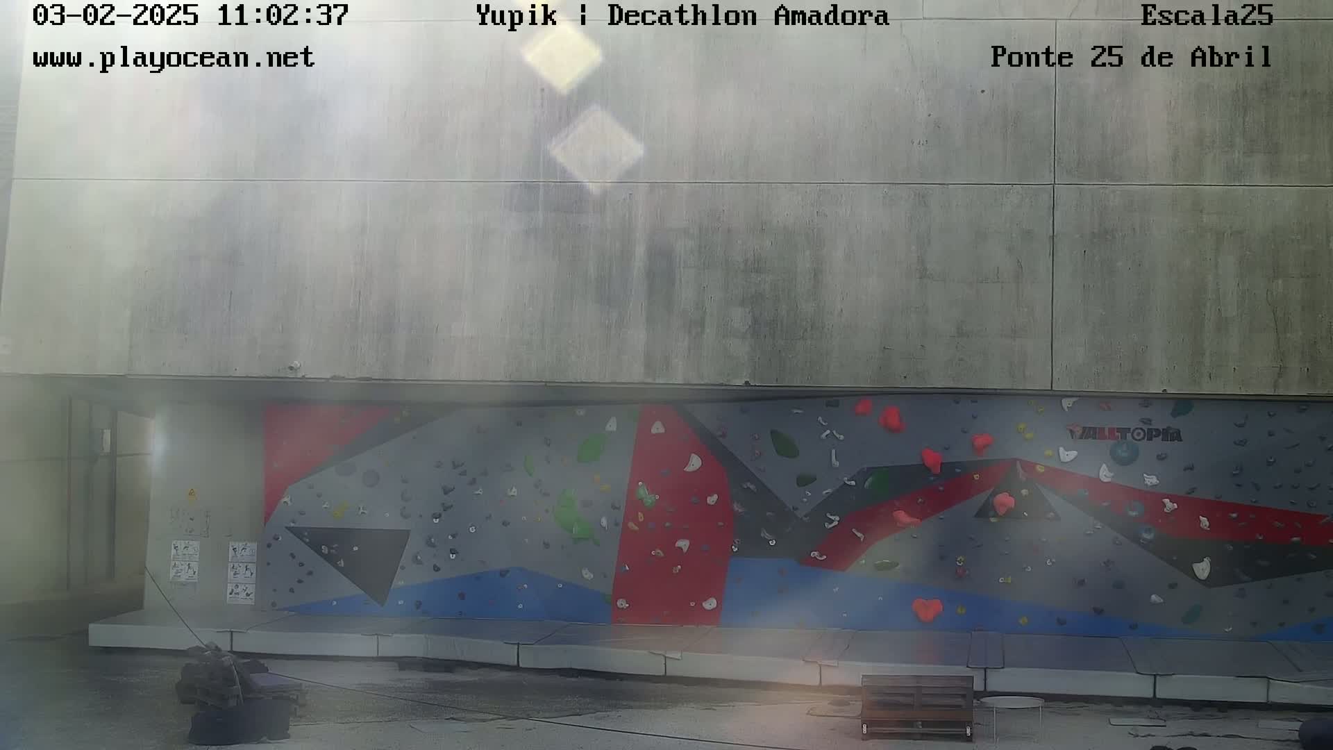An outdoor climbing wall with various colored holds is situated against a large concrete wall under hazy conditions.