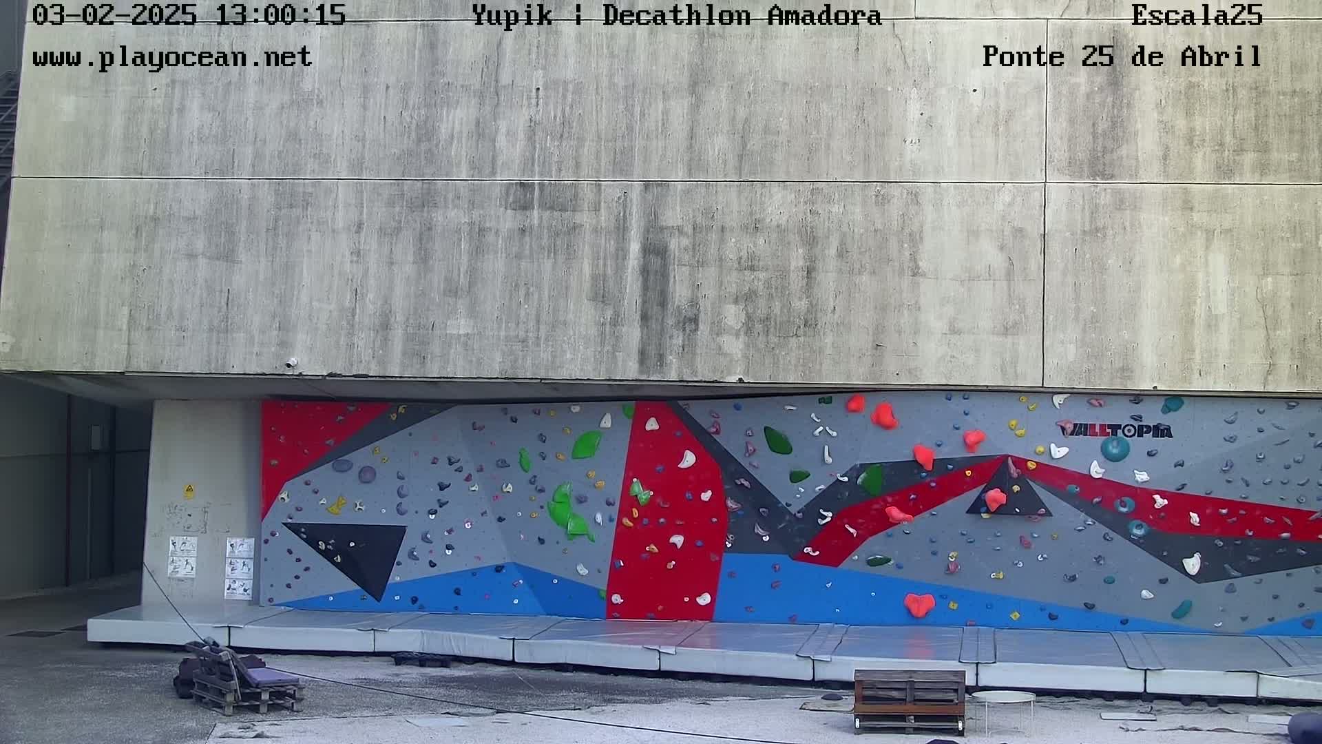 An outdoor climbing wall with various colored holds is situated beneath a large concrete structure on a mostly clear day.