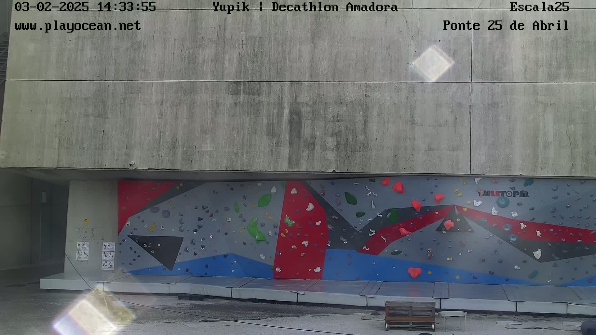 A large outdoor climbing wall with various colored holds is situated beneath a concrete overhang.
