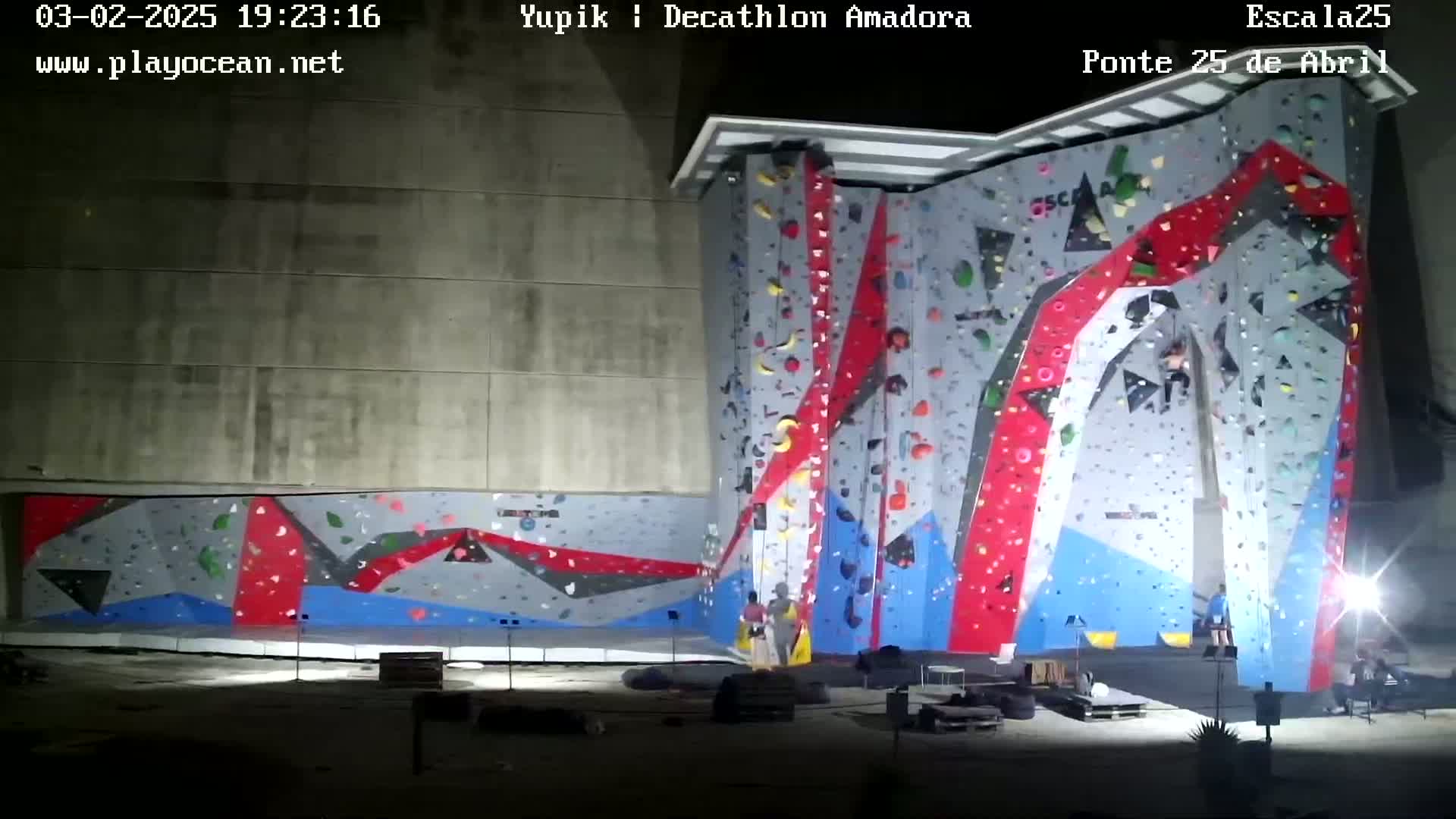 A large, brightly colored climbing wall is illuminated at night, with a few people near the base.