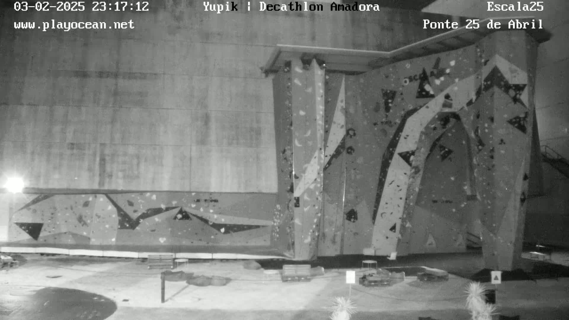 A nighttime black-and-white view of an outdoor climbing wall with various holds and some equipment scattered on the ground in front of it.