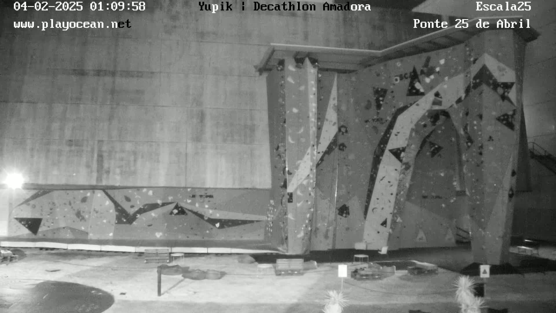A grayscale nighttime image shows an outdoor climbing wall with various holds and geometric designs.