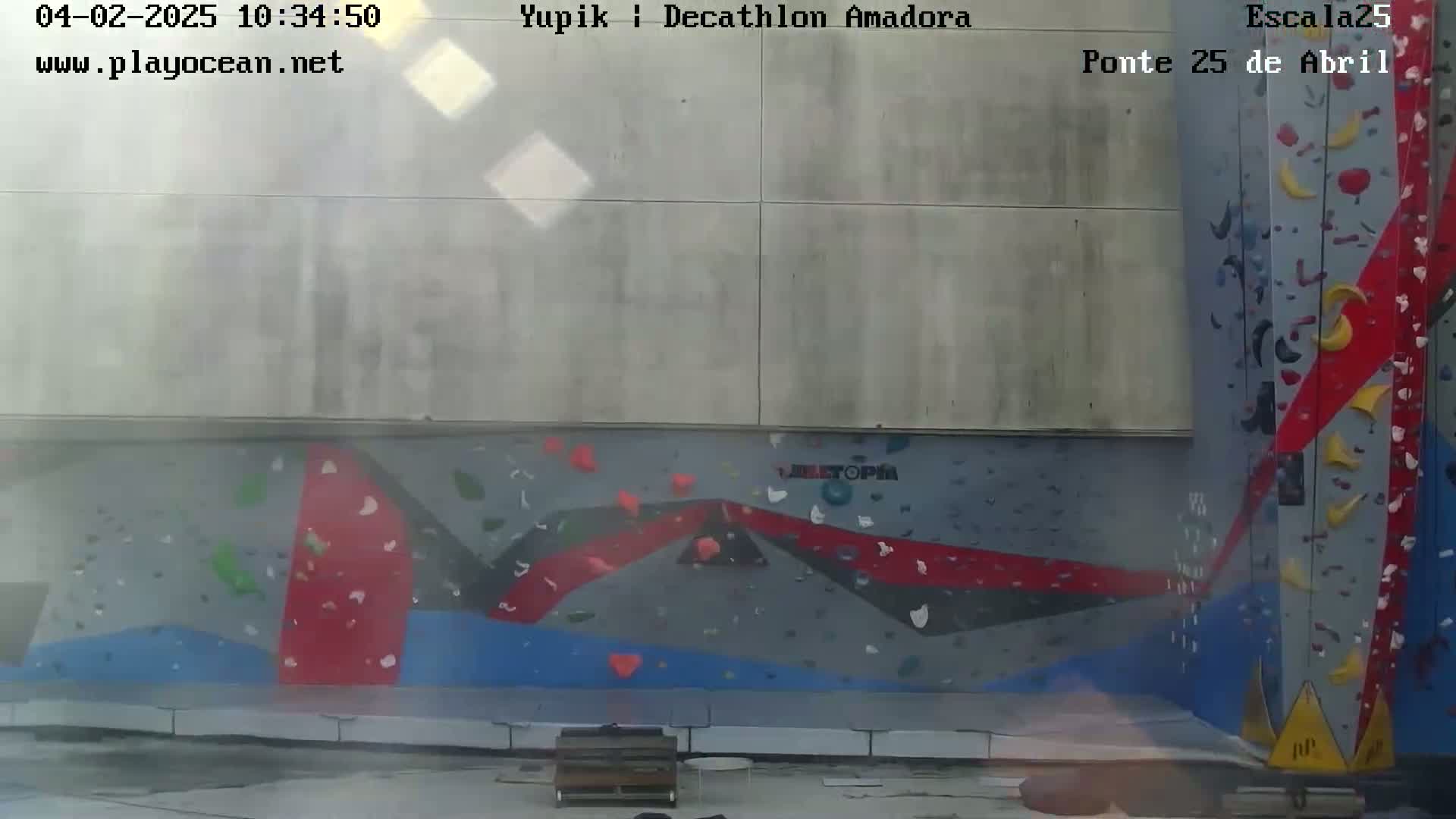 An outdoor view of a large climbing wall with various colored holds, set against a light-grey building under hazy conditions.