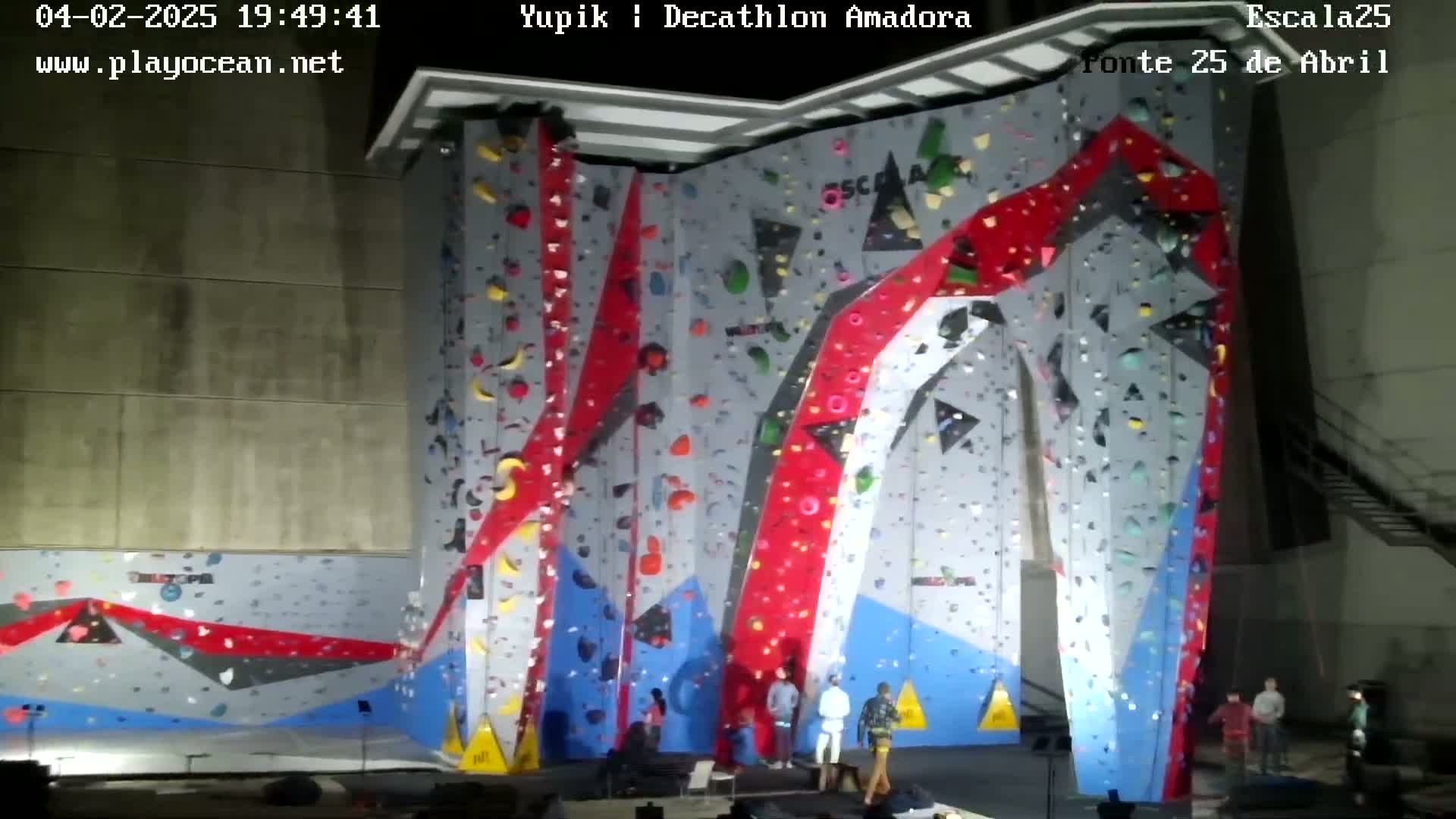 A large indoor climbing wall with various colored holds, and several people standing at the base.