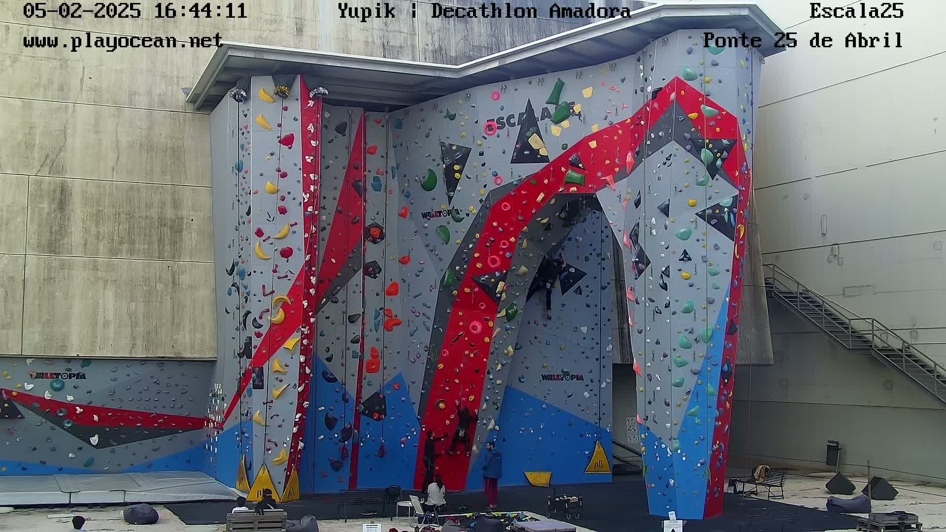 A large, colorful indoor climbing wall with several people climbing and others observing.