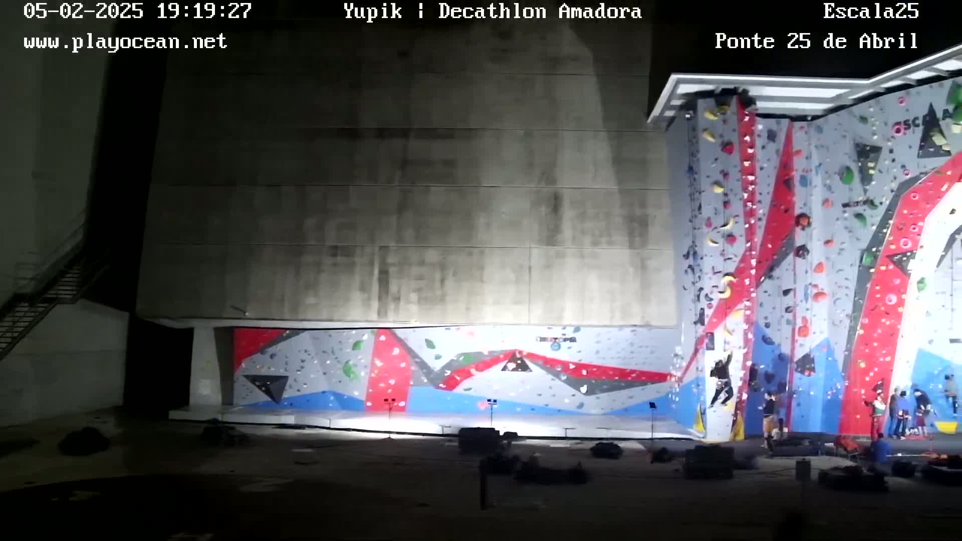 A large, brightly colored indoor climbing wall is illuminated at night, with several climbers ascending it and spectators watching from the base.