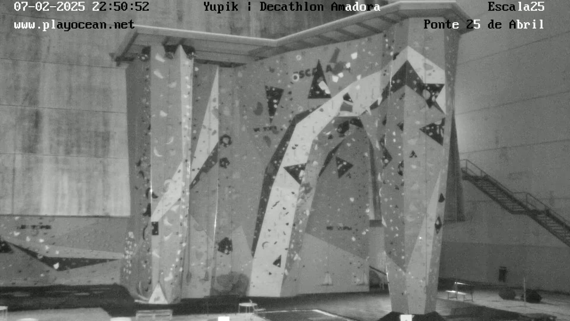 A grayscale image shows an indoor climbing wall with various holds and routes, situated within a large, empty space.