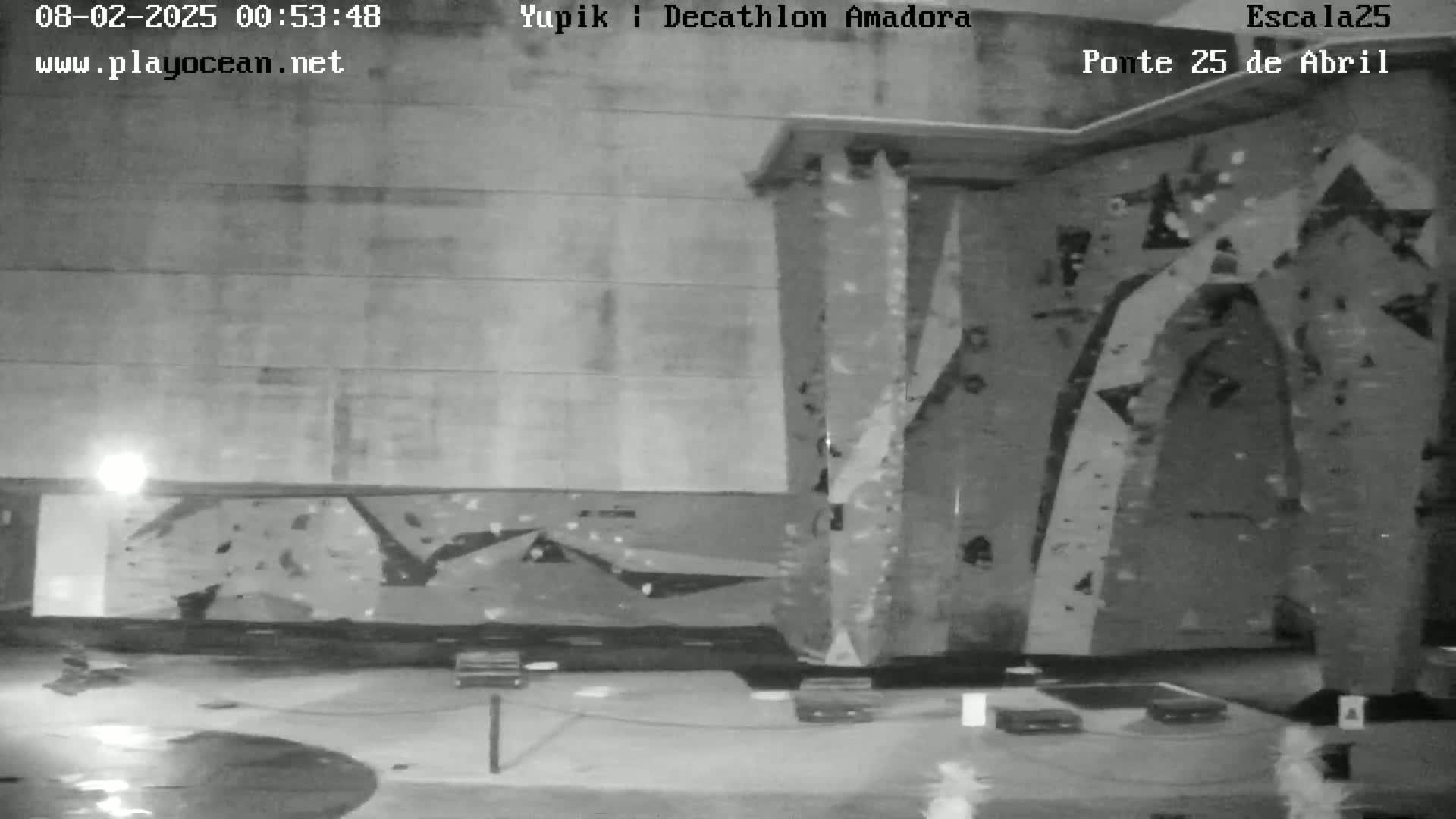 A black and white nighttime image shows an indoor climbing wall with various holds and textured surfaces, and a wet floor.
