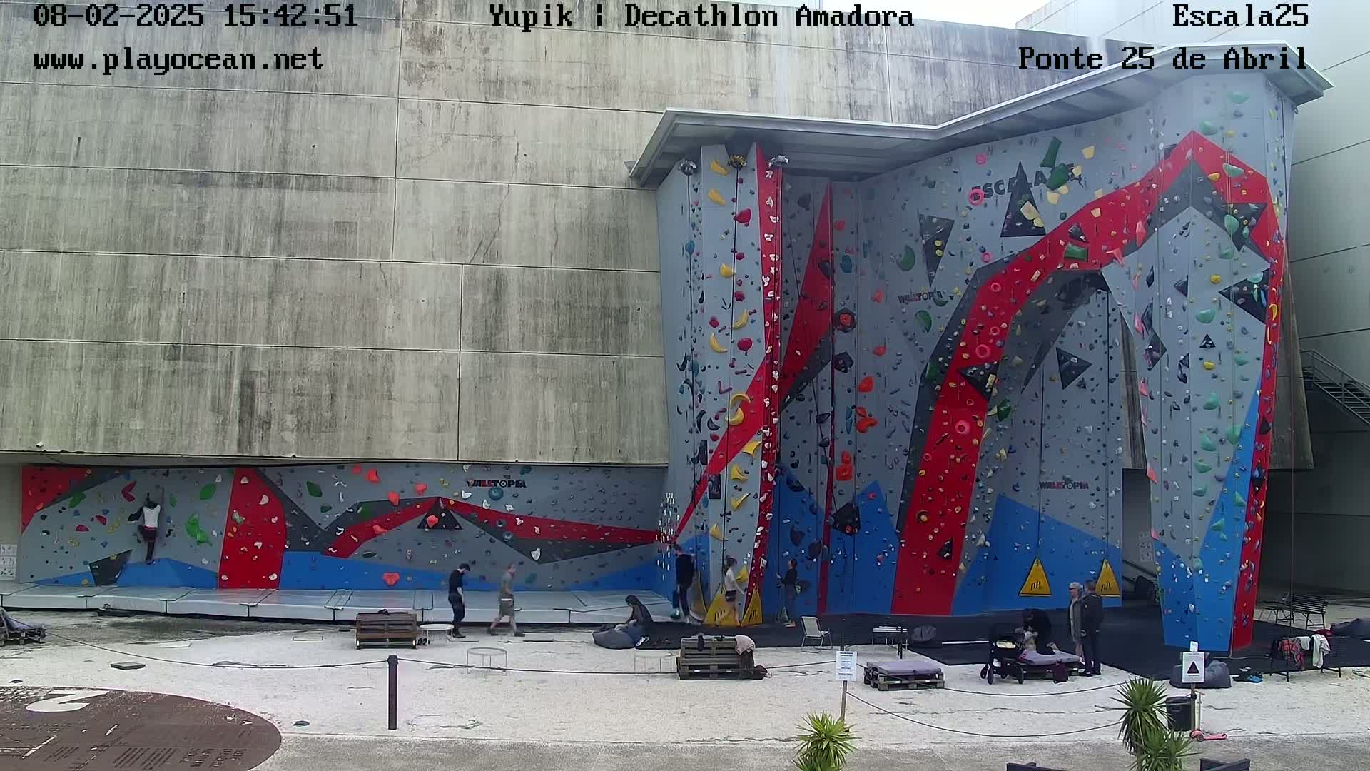An outdoor climbing wall with several people climbing and others observing, under a mostly sunny sky.