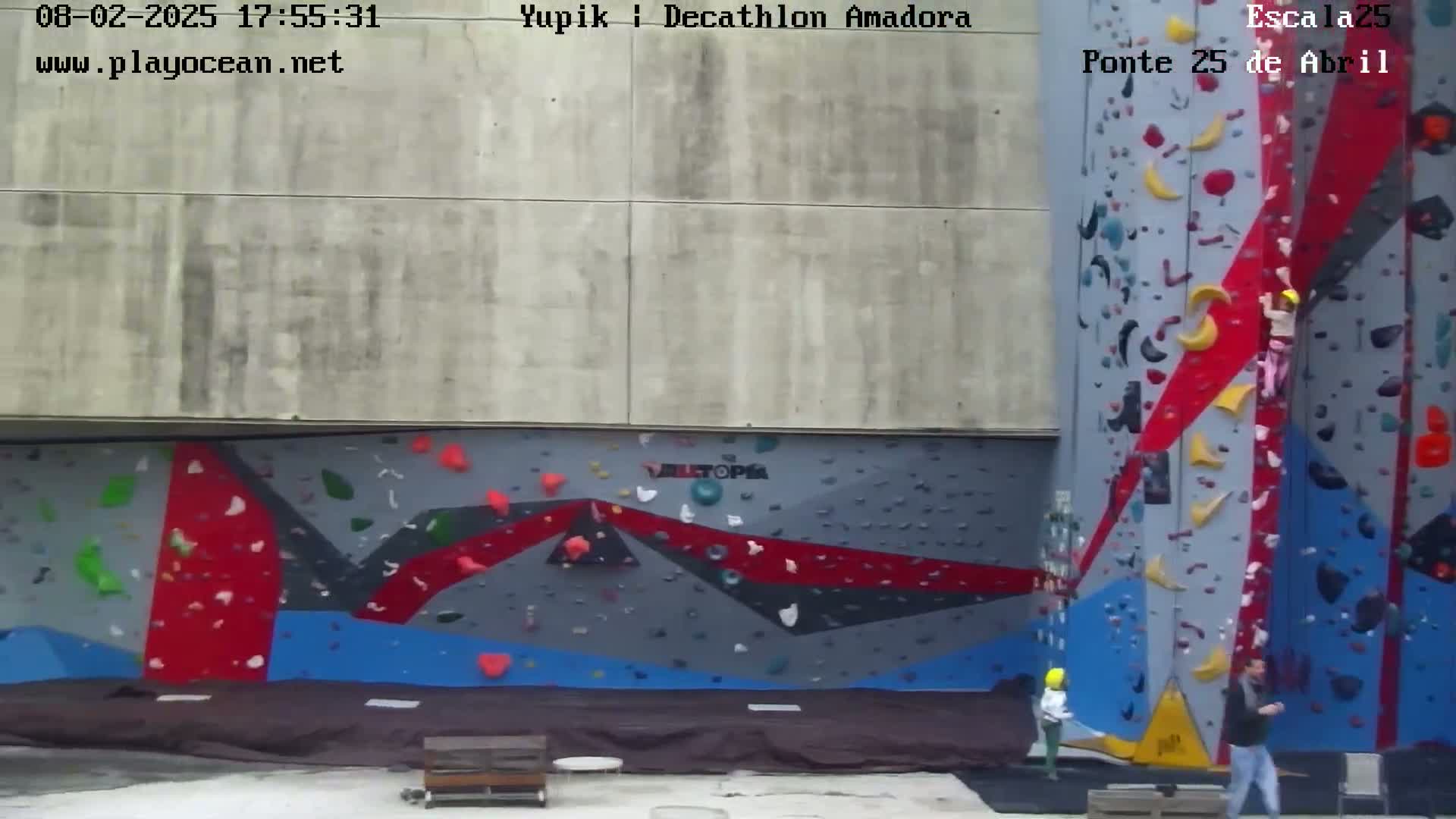 An outdoor climbing wall with several people climbing and watching, under a cloudy sky.