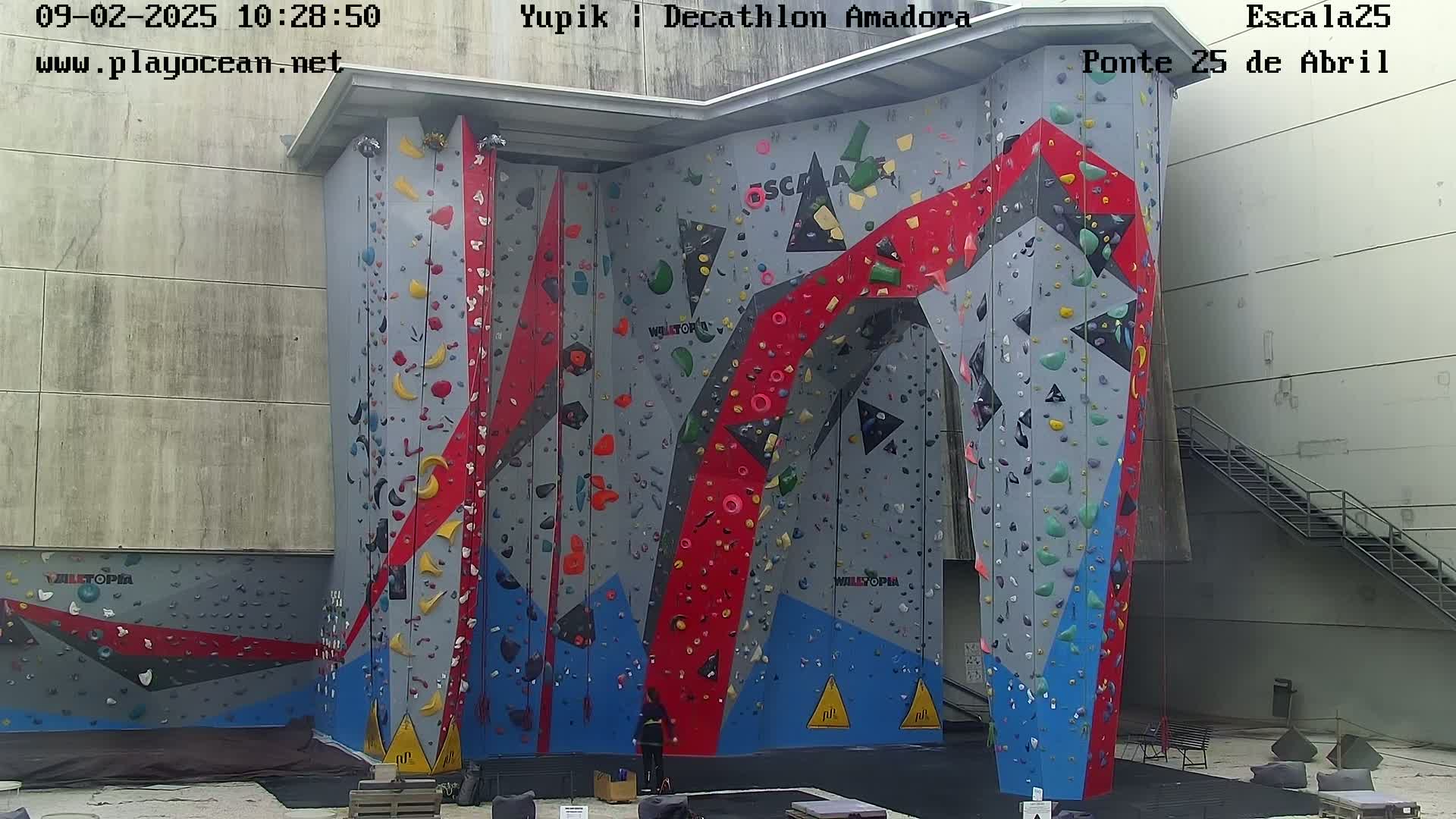 A large indoor climbing wall with various colored holds is seen on a sunny day.