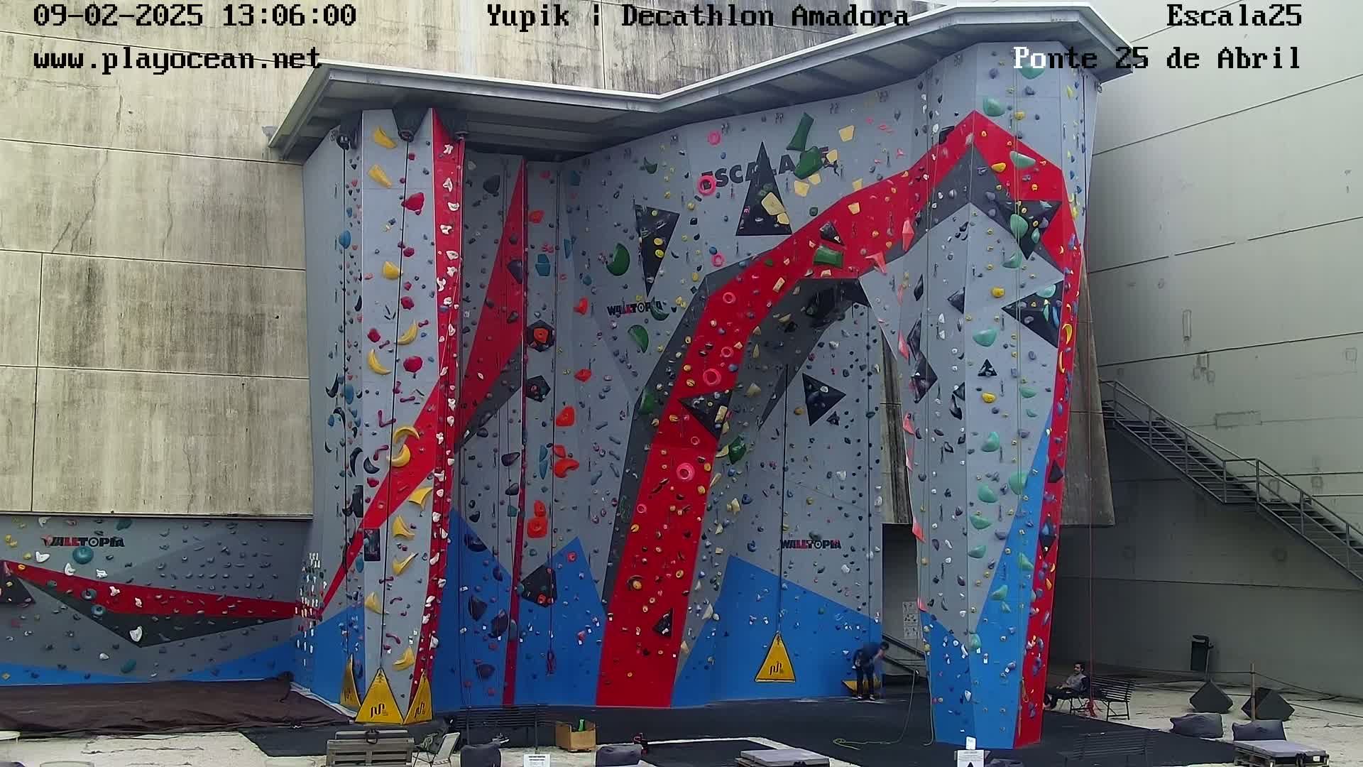 An outdoor climbing wall with various colored holds is situated next to a large gray building.