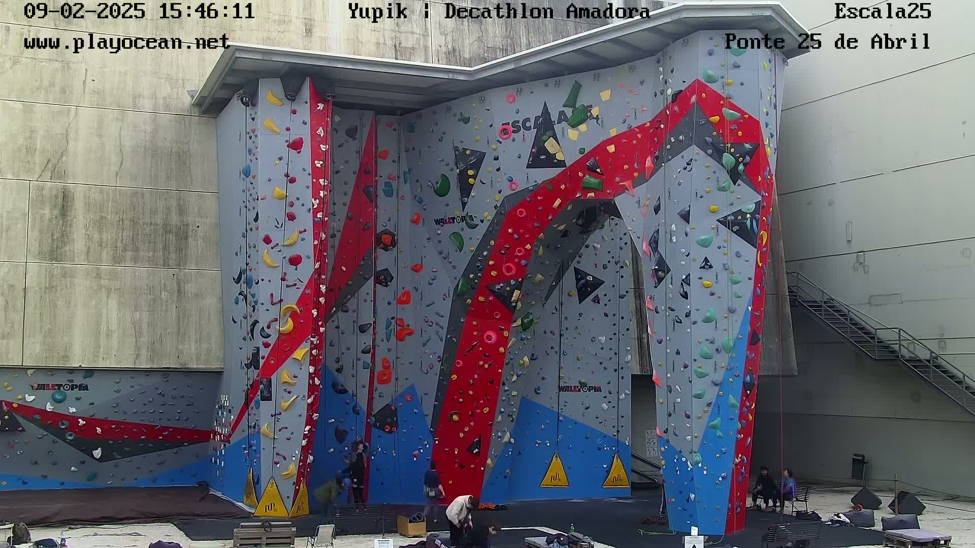 An outdoor climbing wall with various colored holds is situated against a building, with a few people near the base on an overcast day.