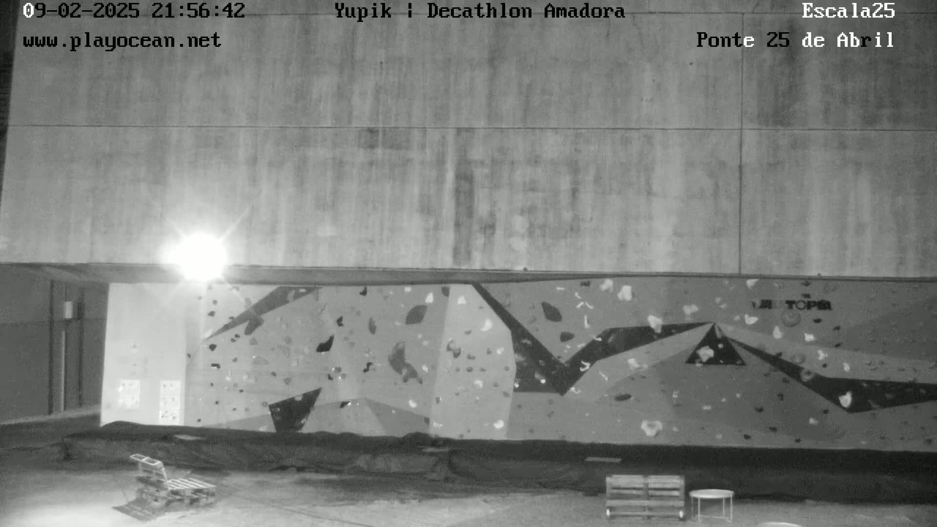 A nighttime, black and white image shows an outdoor climbing wall under a large, concrete overhang.