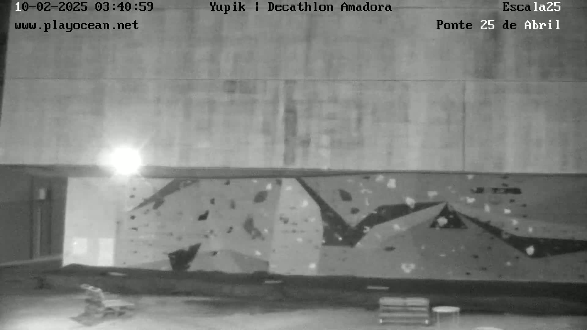 A nighttime, grayscale image shows an indoor climbing wall with sparsely scattered holds under a bright light.
