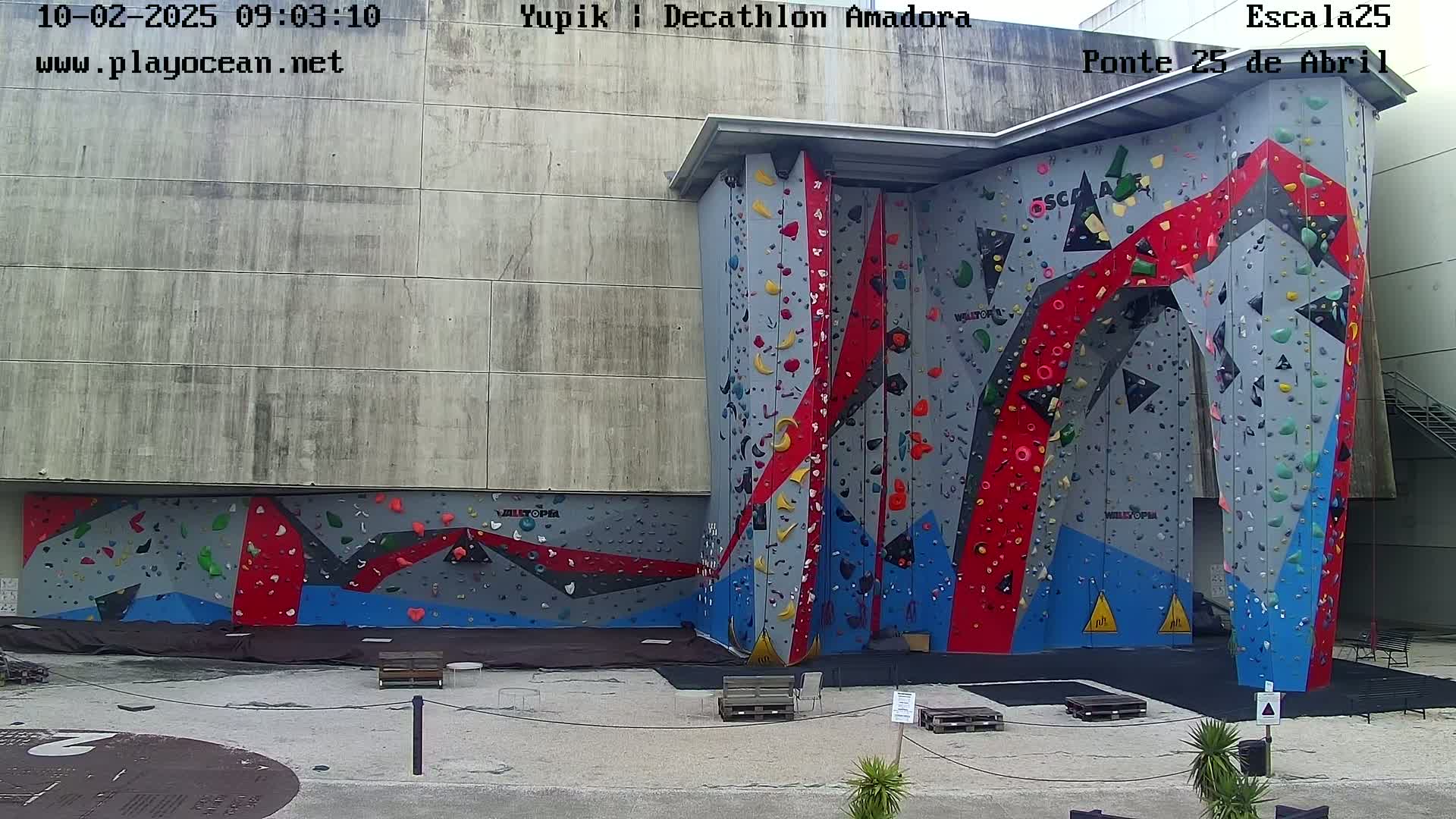 An outdoor climbing wall with red, blue, and gray sections is situated against a concrete building.