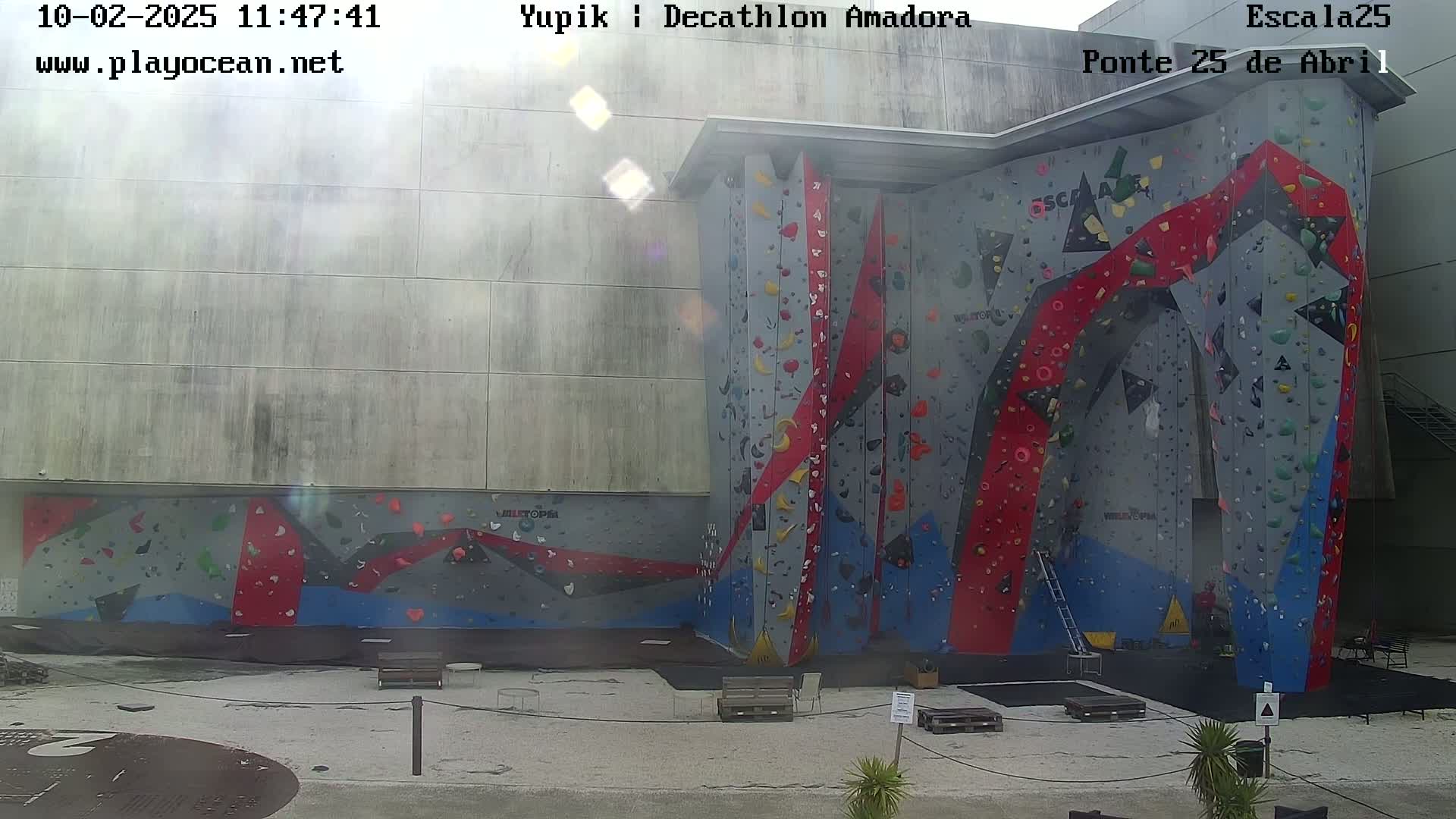 A large outdoor climbing wall with red, gray, and blue sections is partially sunny.