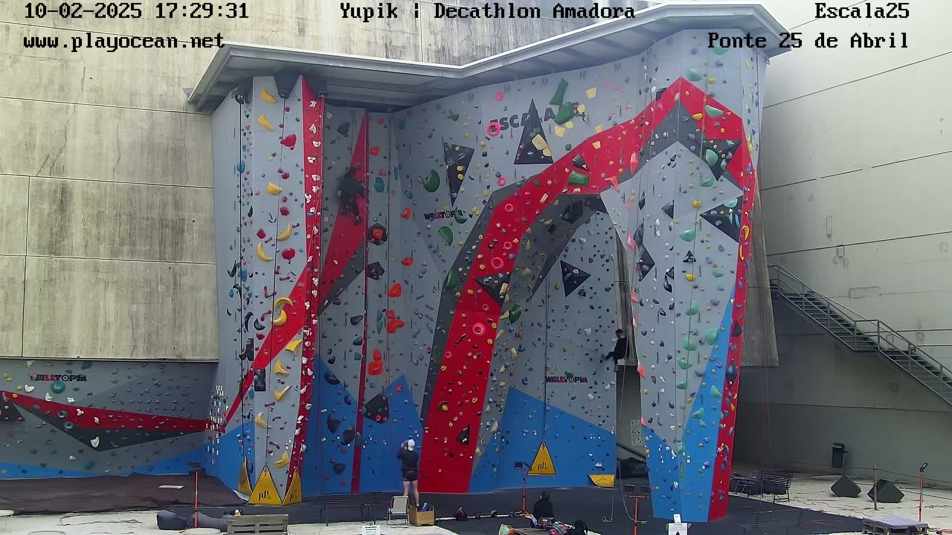A large, colorful indoor climbing wall with several people climbing and others observing, outdoors under overcast skies.