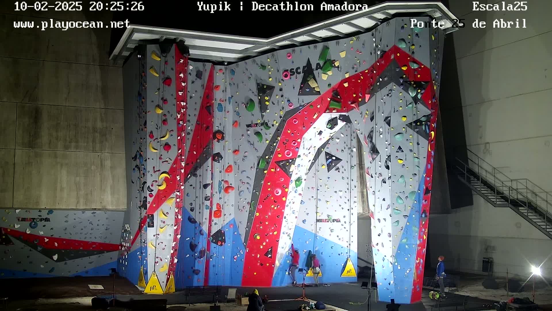 A large indoor climbing wall with various colored holds is illuminated, and a few people are at the base of the wall.