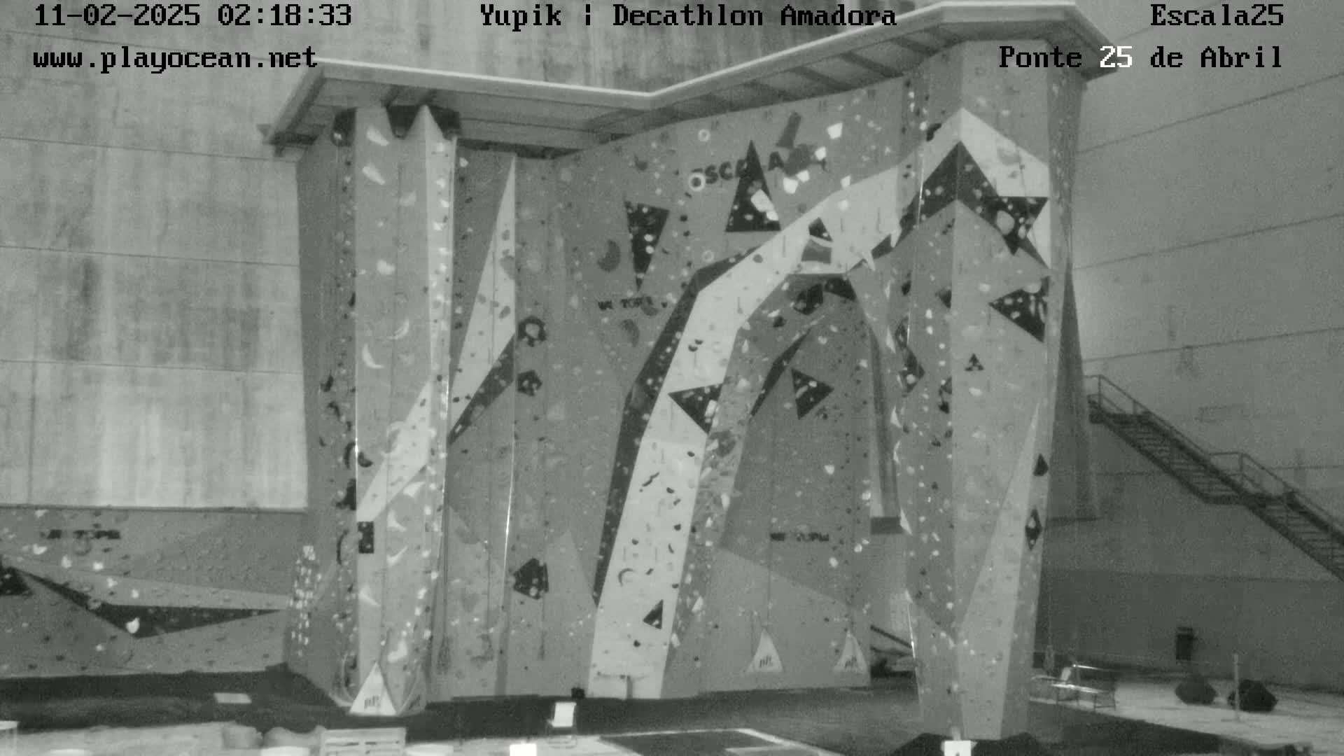 A grayscale image shows an indoor climbing wall with various holds and geometric designs against a concrete wall.
