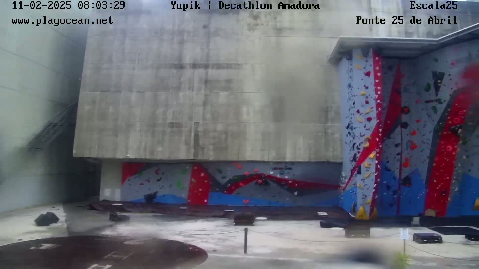 A large outdoor climbing wall with red, blue, and gray holds is situated against a concrete building on a wet, overcast day.