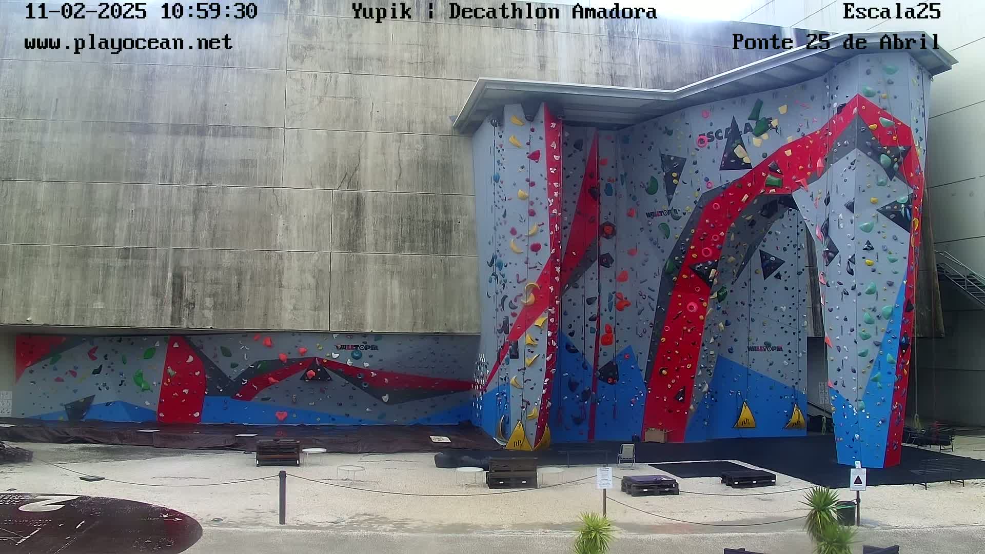 An outdoor climbing wall with various colored holds is situated against a large, gray concrete building on a partly cloudy day.