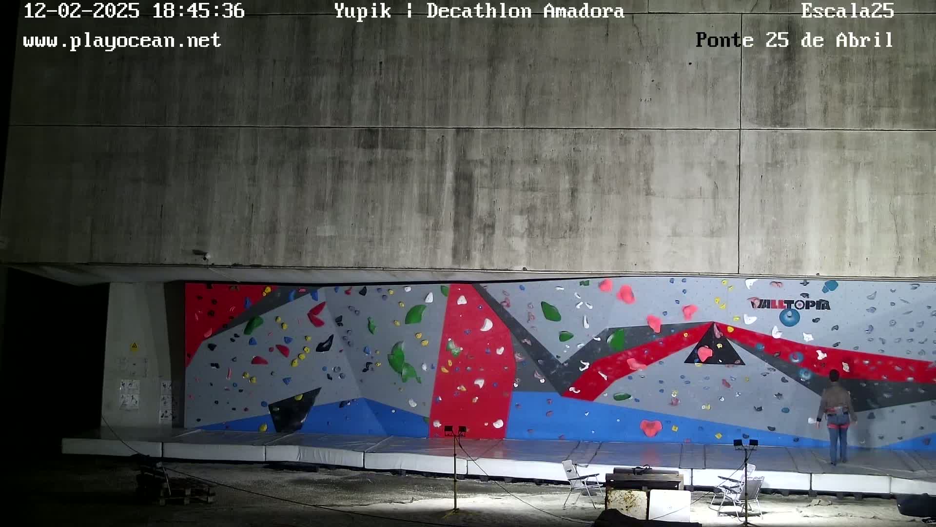 An outdoor nighttime view of a colorful climbing wall with two people standing at its base.