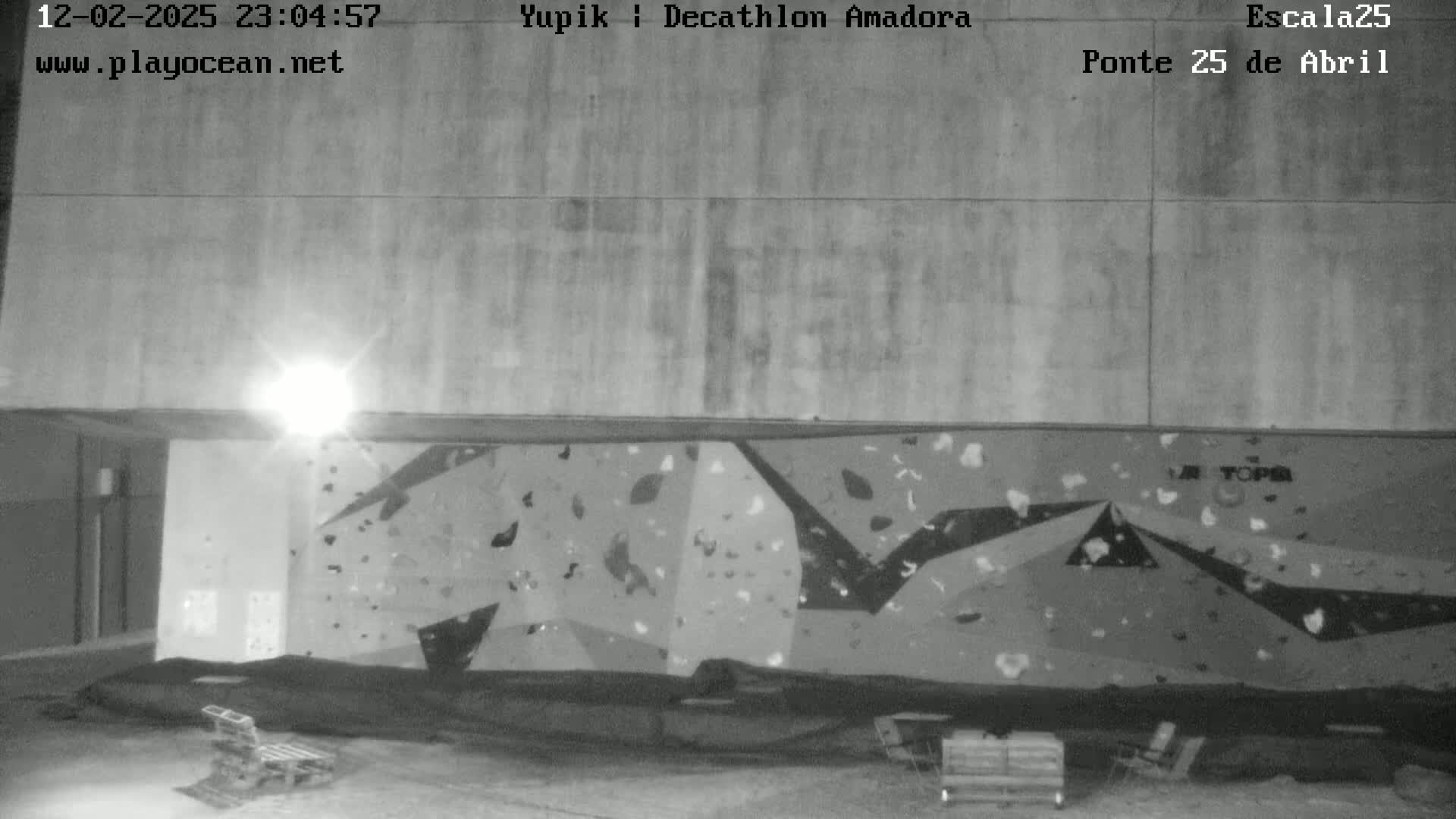 A nighttime, black-and-white image shows an outdoor climbing wall under a light source, with some benches and other equipment nearby.