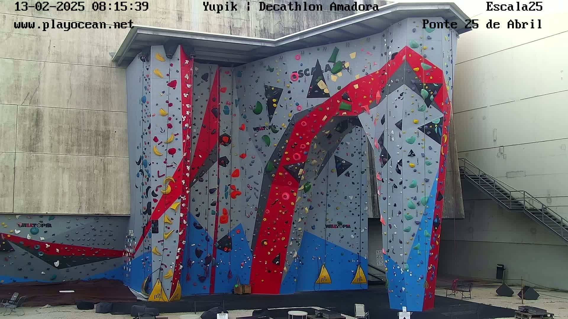 A large, multicolored indoor climbing wall stands against a light gray building, next to a metal staircase.