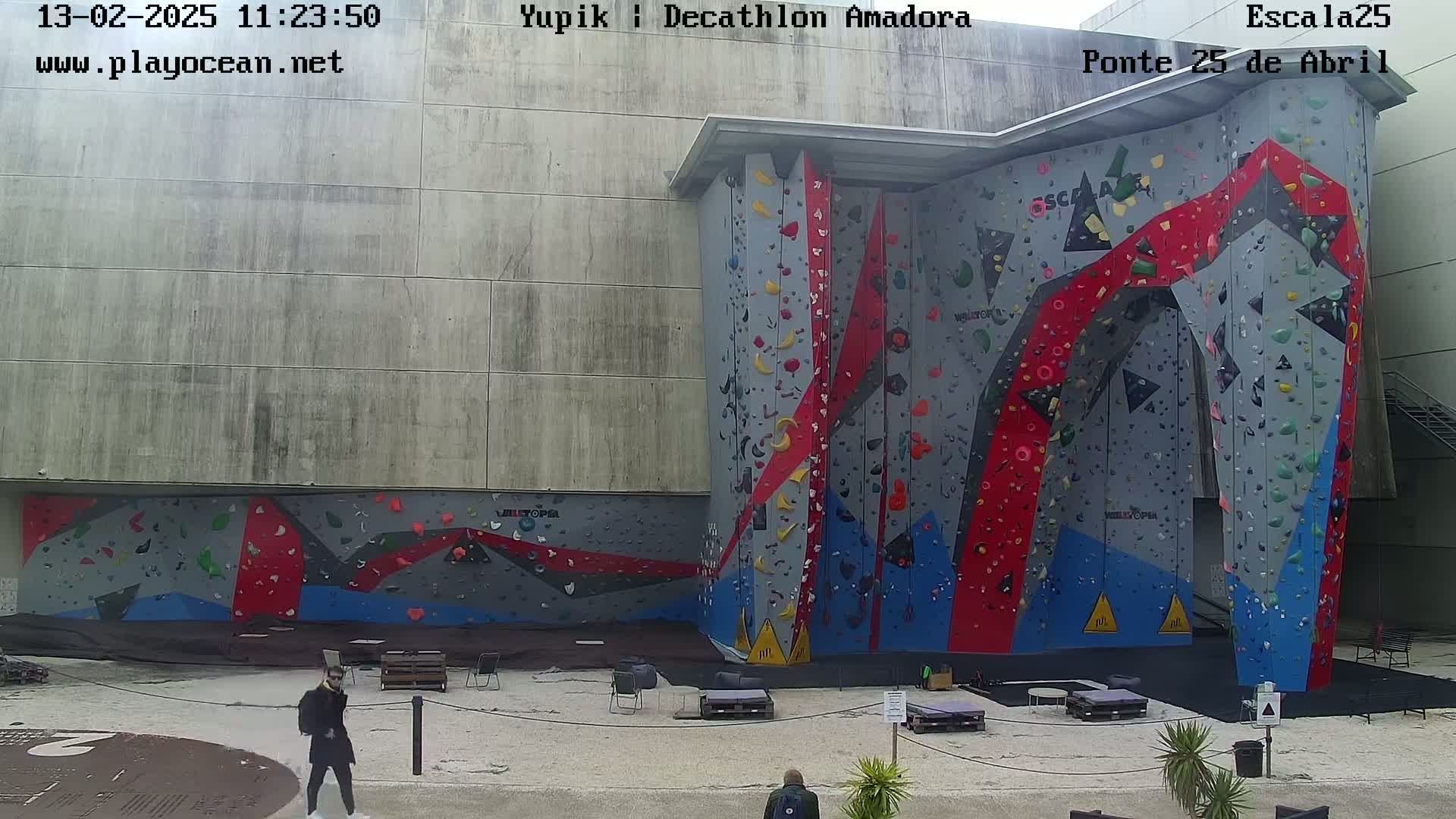 A large outdoor climbing wall with red, blue, and gray holds is situated against a concrete building on a partly sunny day.