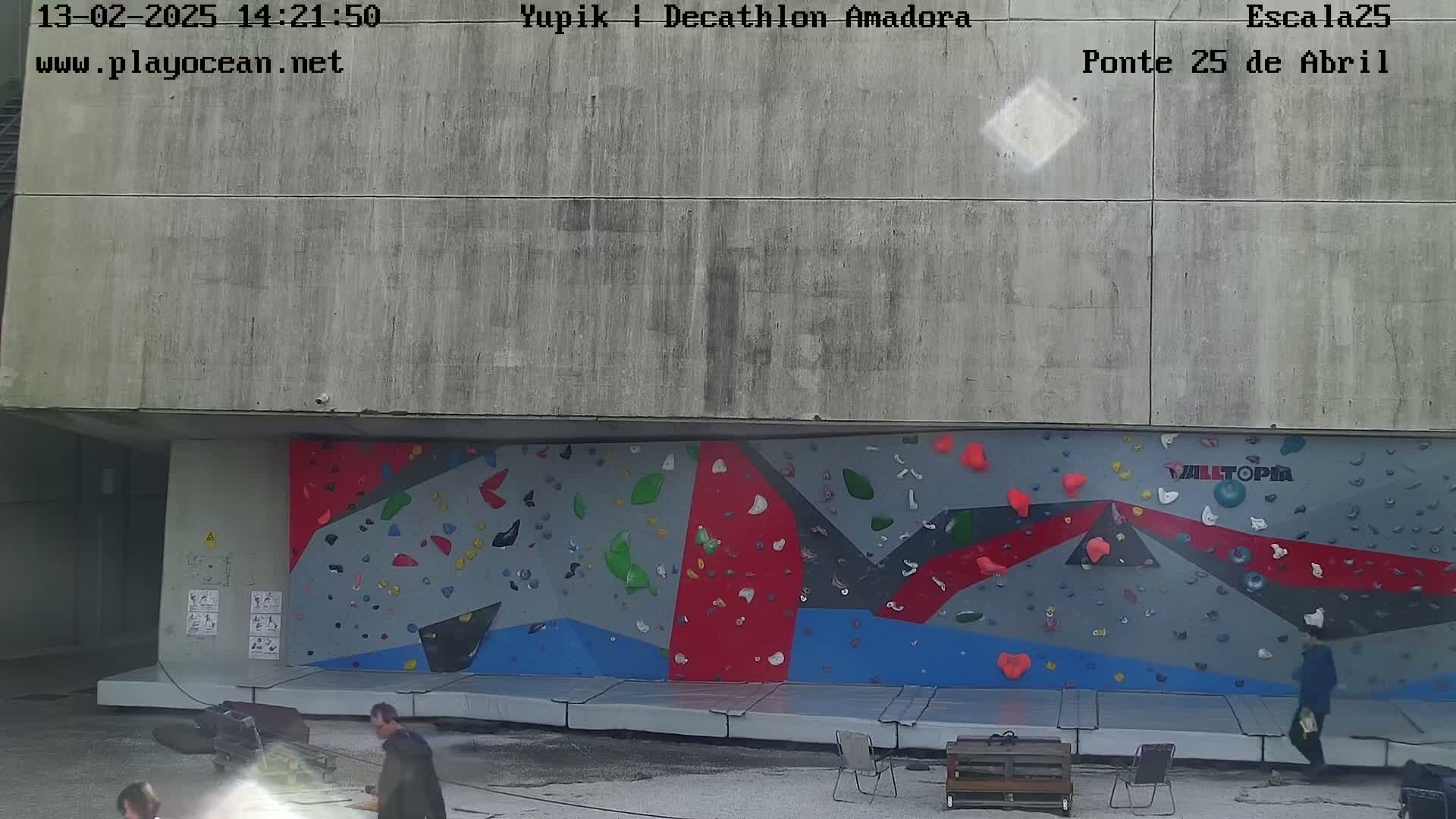 An outdoor climbing wall with various colored holds, situated beneath a concrete overhang, is partially surrounded by people and some outdoor furniture on a mostly clear day.
