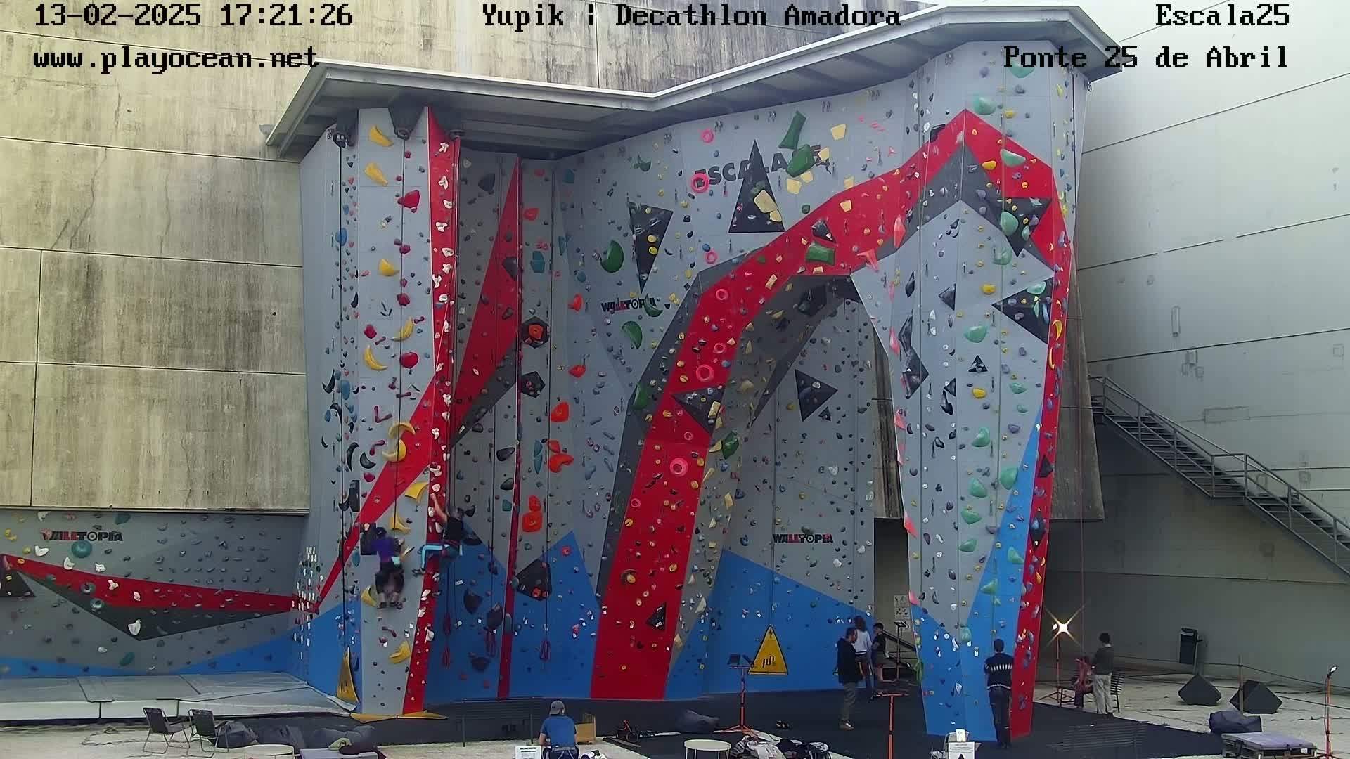 A large, colorful indoor climbing wall with several people climbing and others watching, located outdoors on a partly cloudy day.